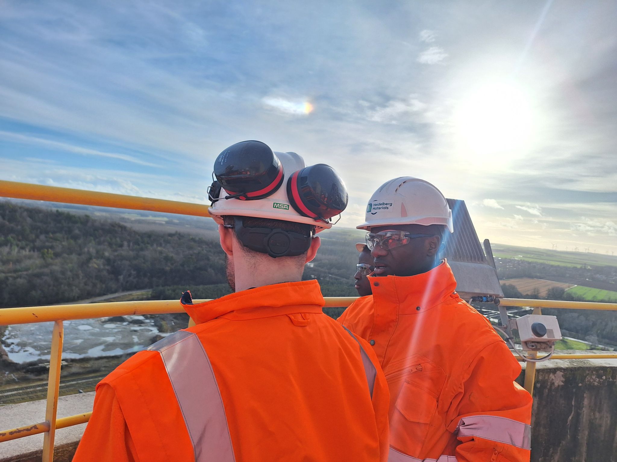 Deux personnes en tenue de sécurité orange et casques blancs se tiennent sur une plateforme élevée, regardant un paysage rural avec collines et soleil lumineux en arrière-plan. Des équipements industriels sont visibles à proximité.