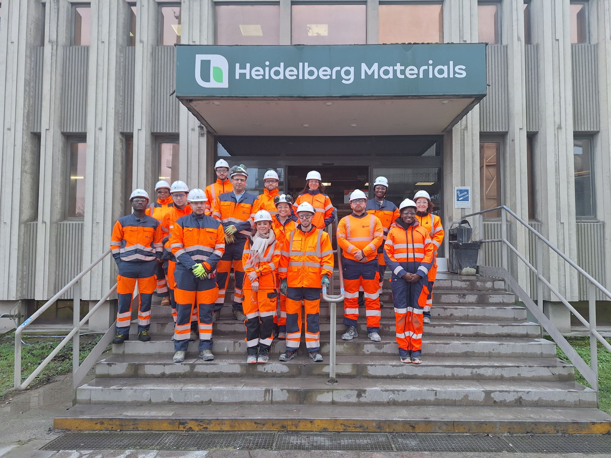 Groupe de travailleurs en tenue de sécurité orange et casques, debout sur des marches devant un bâtiment marqué "Heidelberg Materials".