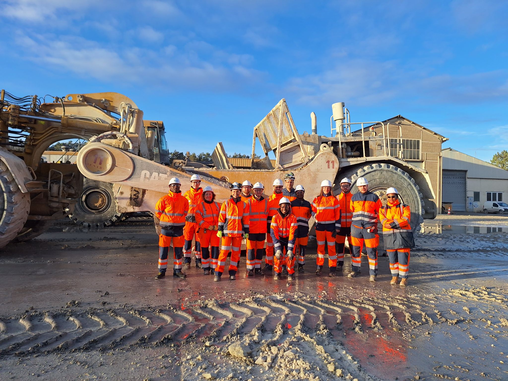 Un groupe de travailleurs en tenue de sécurité orange se tient devant une grande machine industrielle dans un environnement extérieur, avec un bâtiment en arrière-plan sous un ciel ensoleillé.