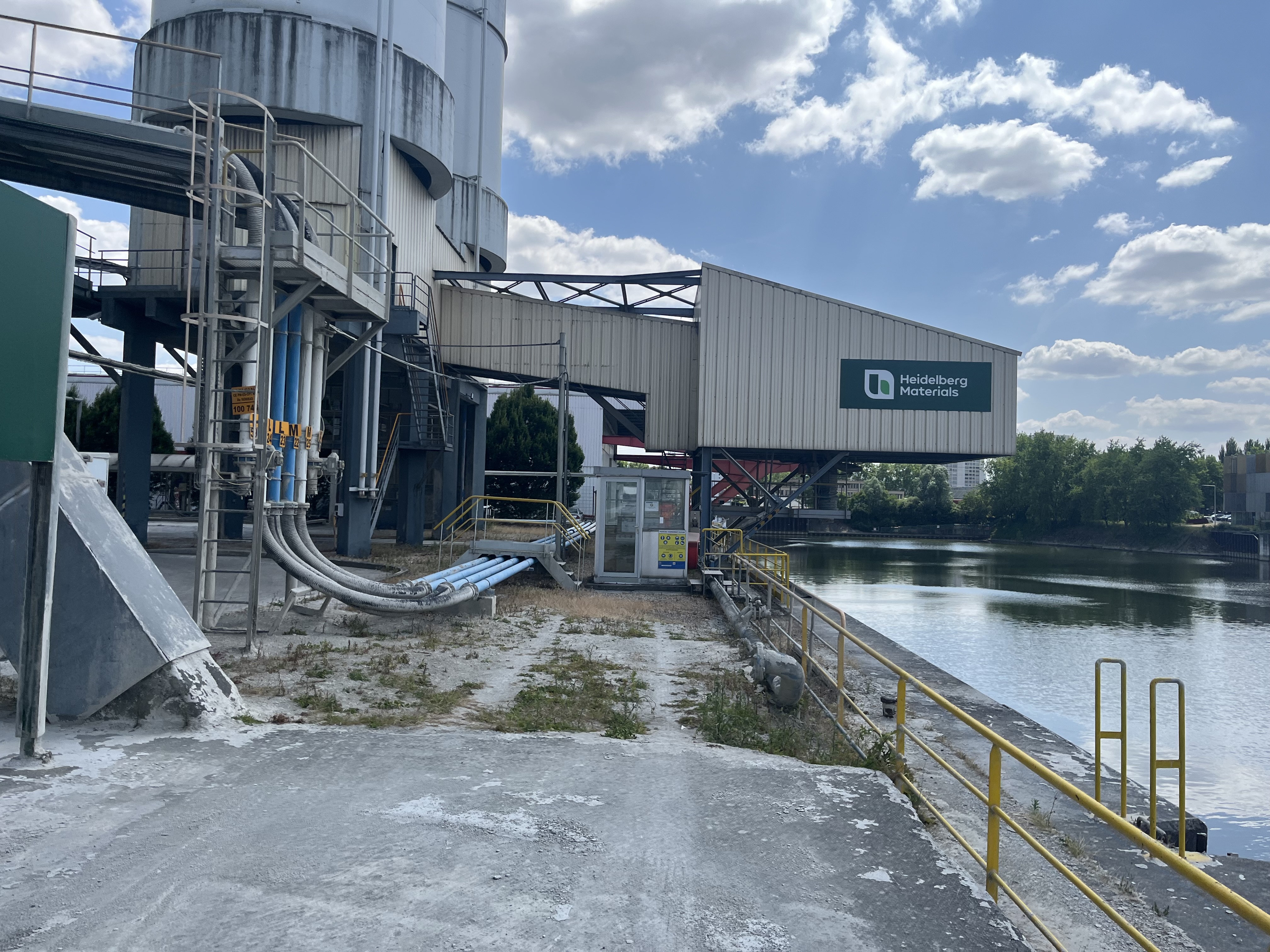 Installation industrielle à côté d'une voie navigable sous un ciel partiellement nuageux. La structure comprend de grands silos à gauche, un système de convoyage s'étendant vers un bâtiment avec un panneau vert et des garde-corps de sécurité bordant l'eau à droite.