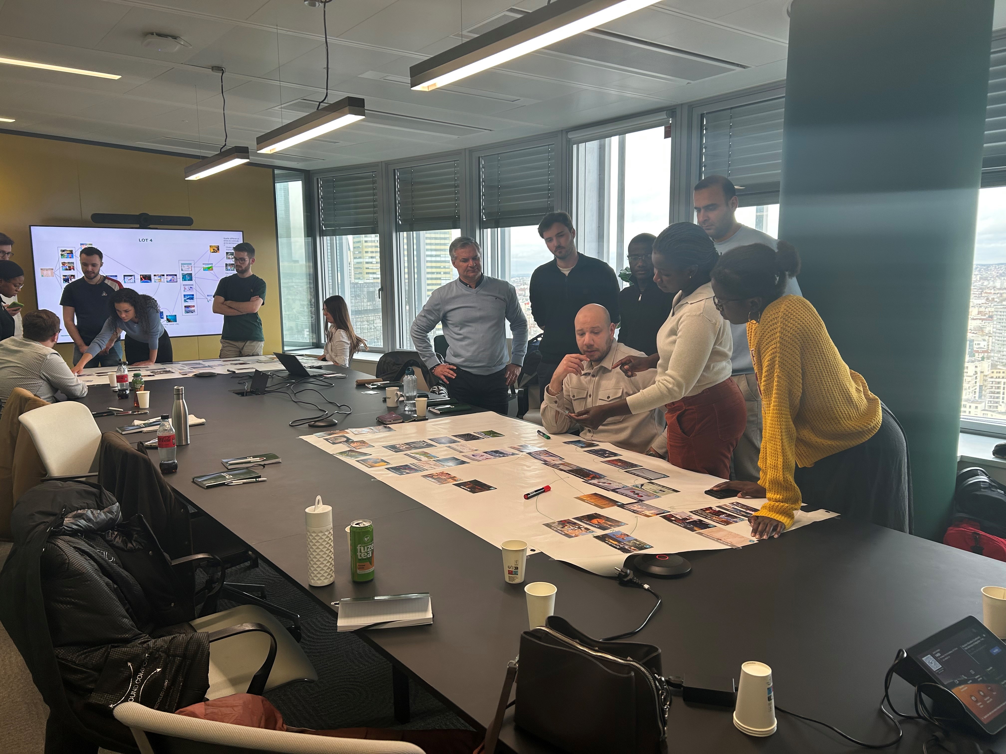Un groupe de personnes se tient autour d'une grande table dans une salle de réunion, examinant des photos et documents étalés. Un écran est visible à gauche avec des graphiques ou des éléments numériques. L'ambiance semble collaborative.
