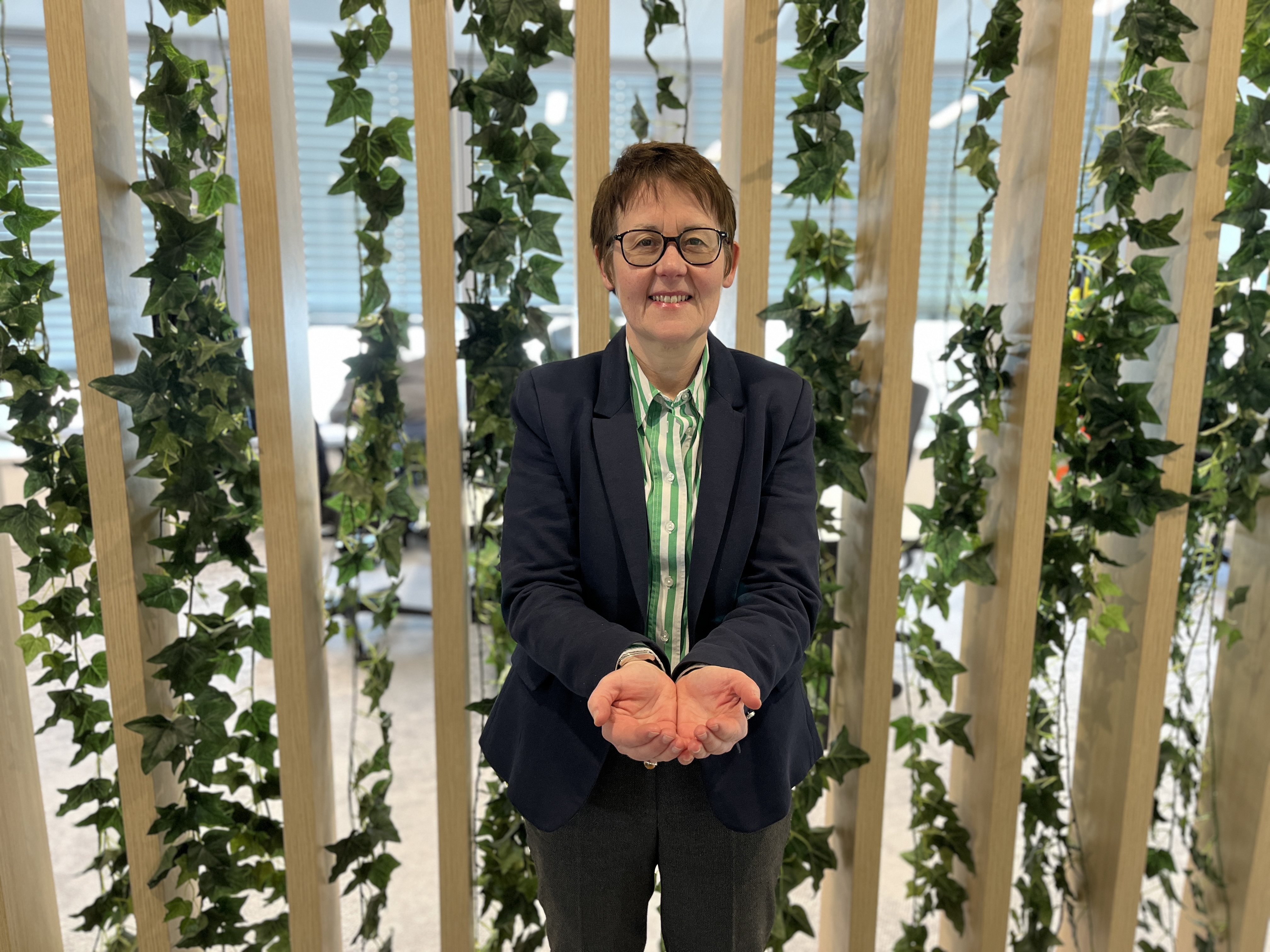 A person in a suit is holding their hands out in front of a vertical wooden structure with green ivy.