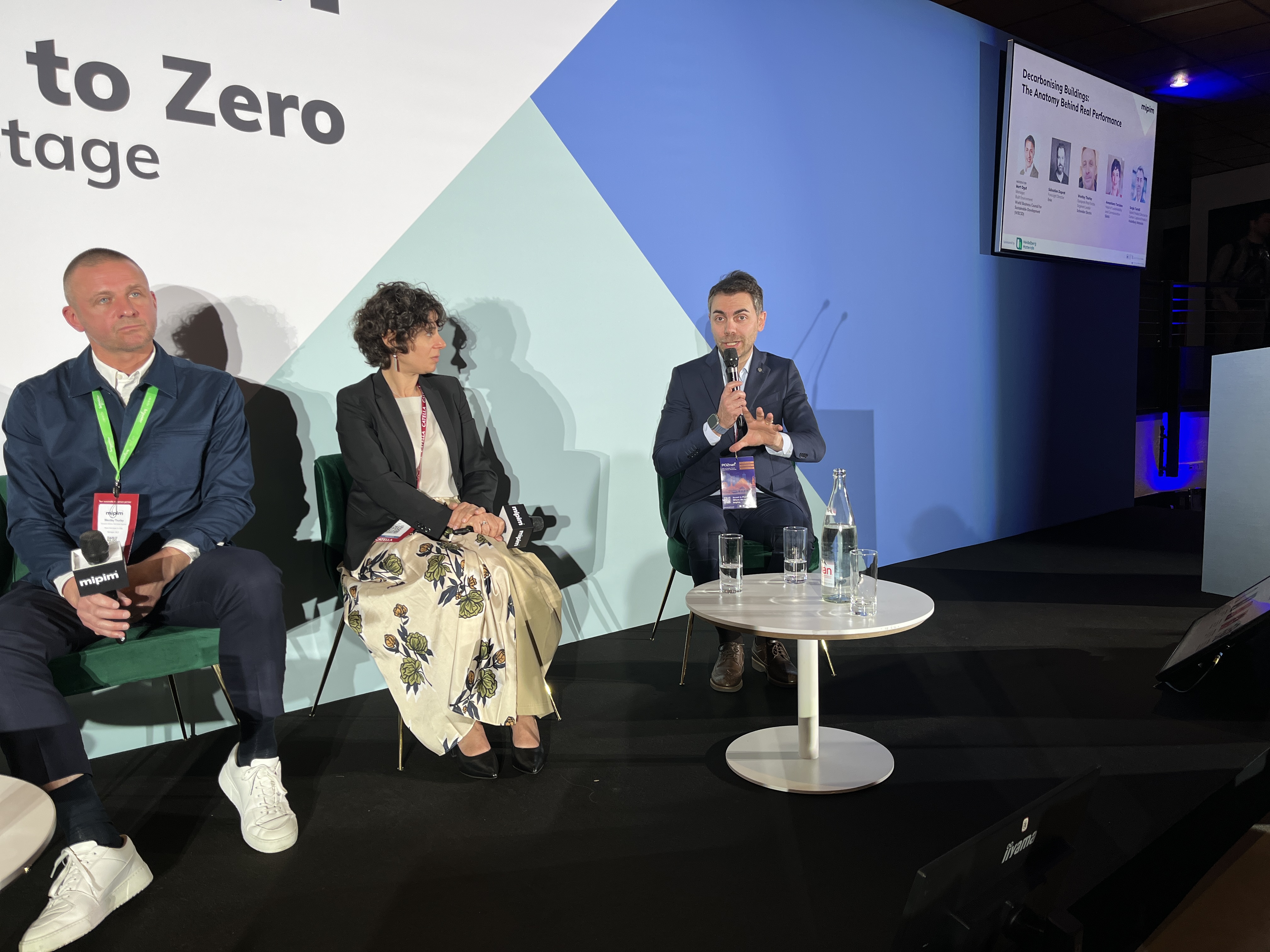 Des intervenants assis sur une scène lors d'une conférence, avec un écran affichant des informations en arrière-plan.