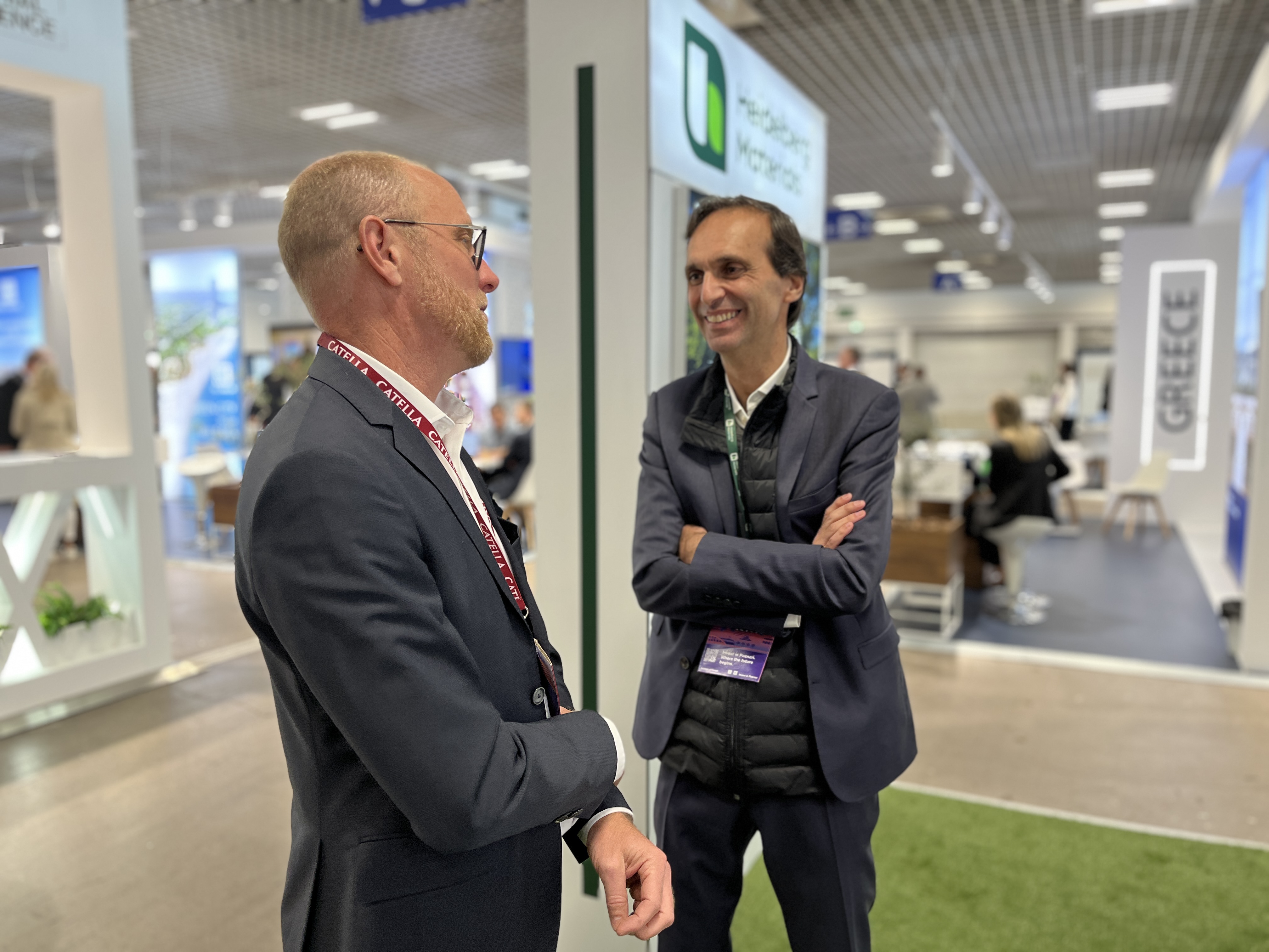 Deux personnes discutent dans un espace d'exposition industriel avec des stands et des panneaux visibles en arrière-plan.
