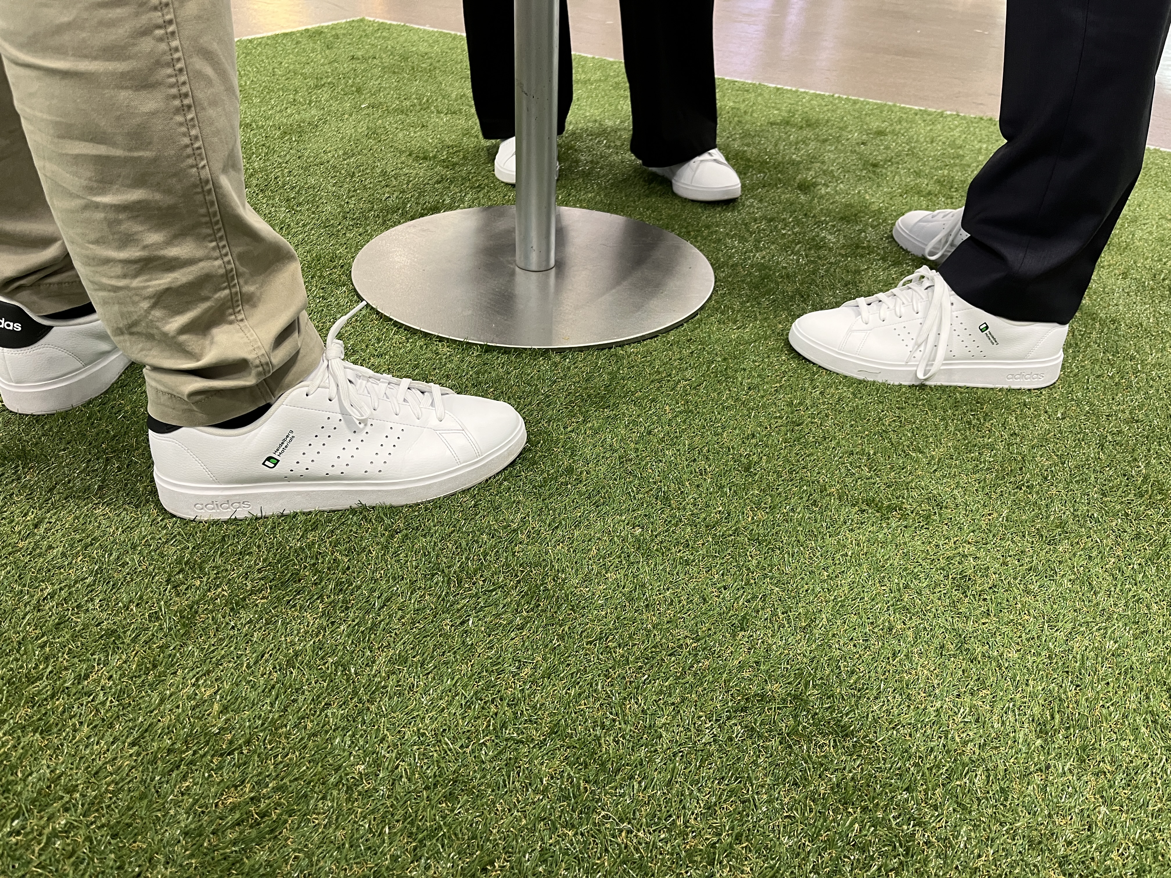 Des personnes debout sur une surface en gazon synthétique, portant des chaussures blanches autour d'une table.