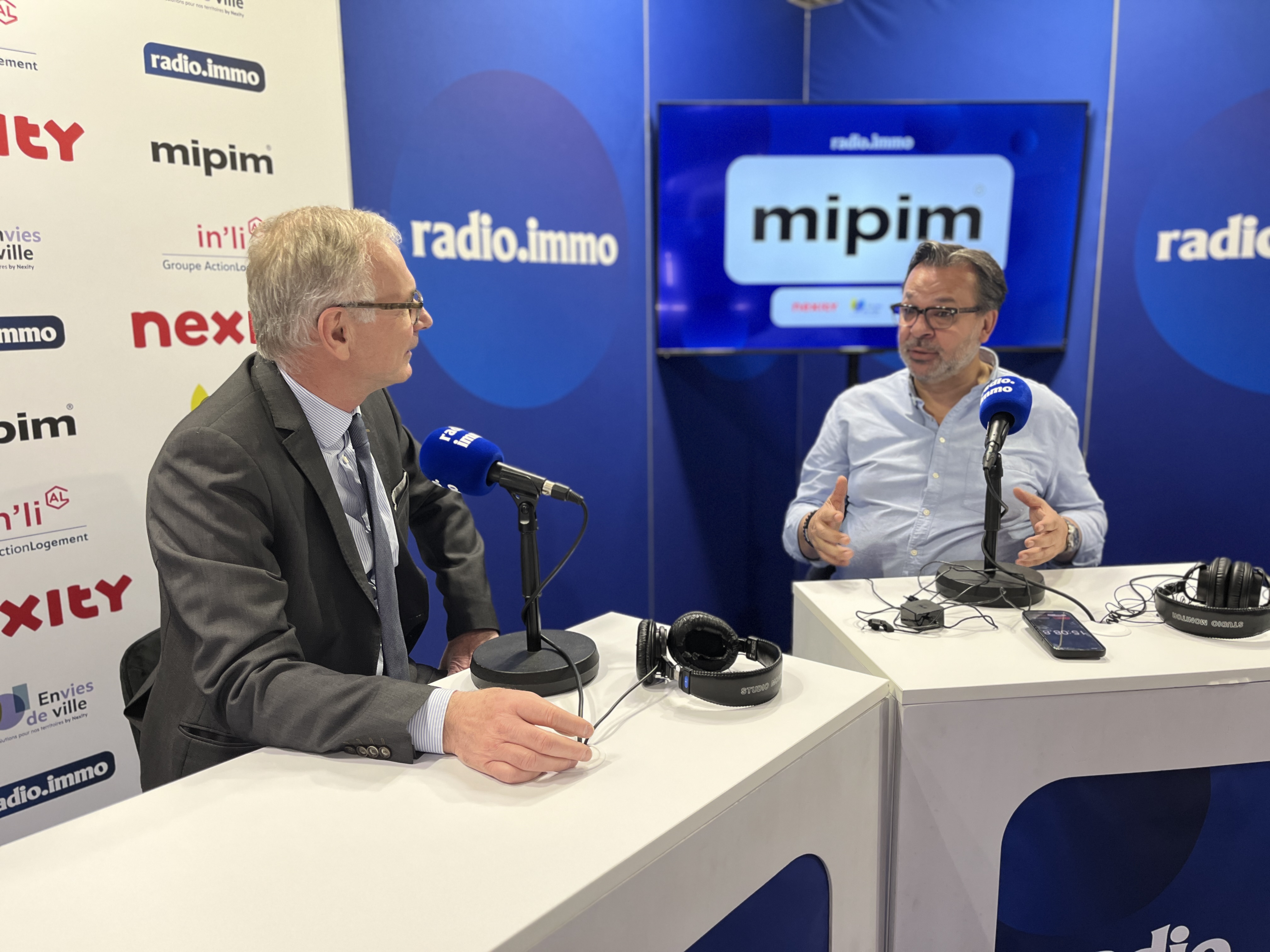 Deux hommes discutent lors d'une émission enregistrée dans un studio à MIPIM, avec des microphones et un écran en arrière-plan.