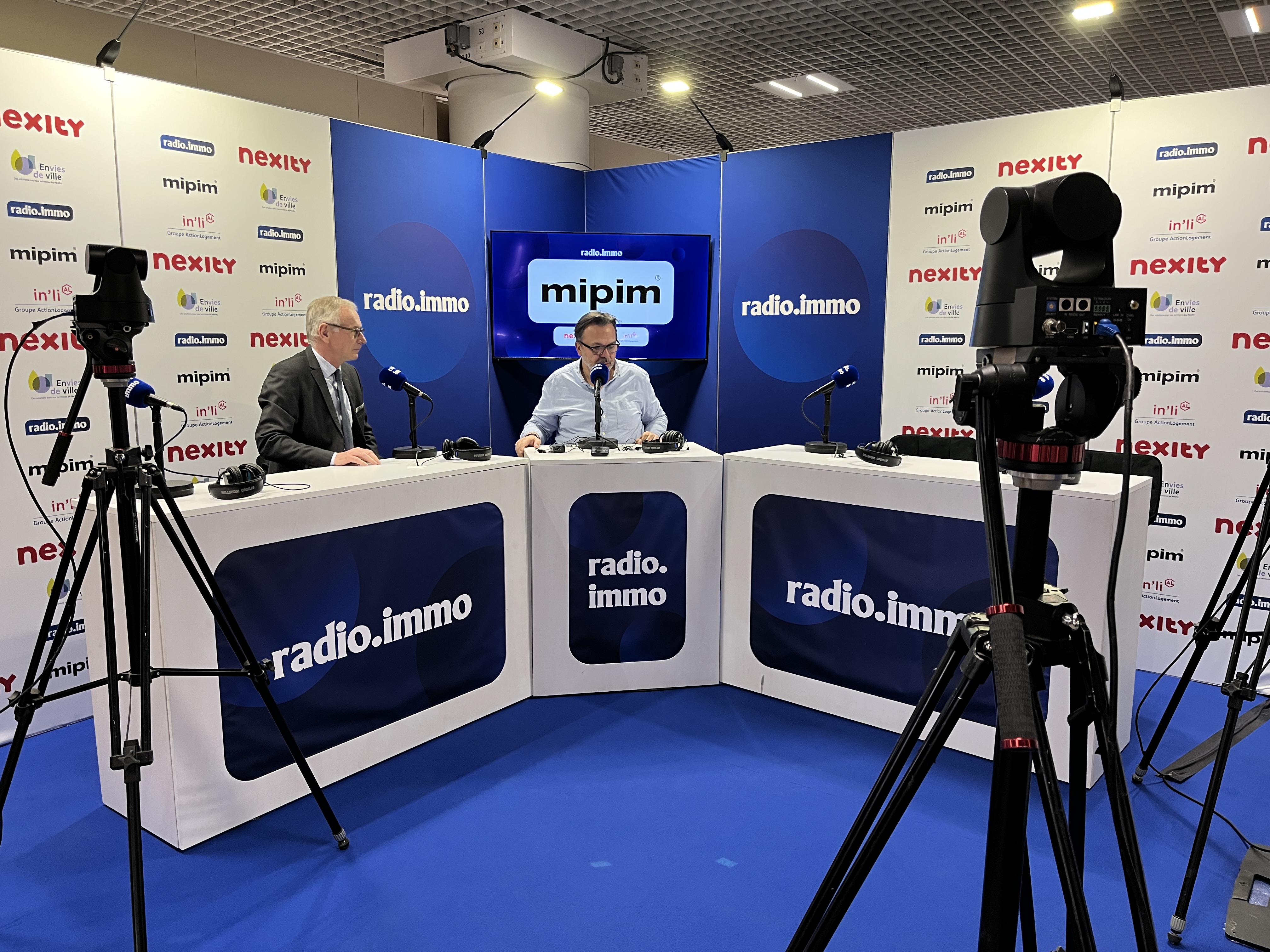 Deux hommes discutent dans un studio d’enregistrement sur le thème de l’immobilier, avec des caméras et des micros visibles.