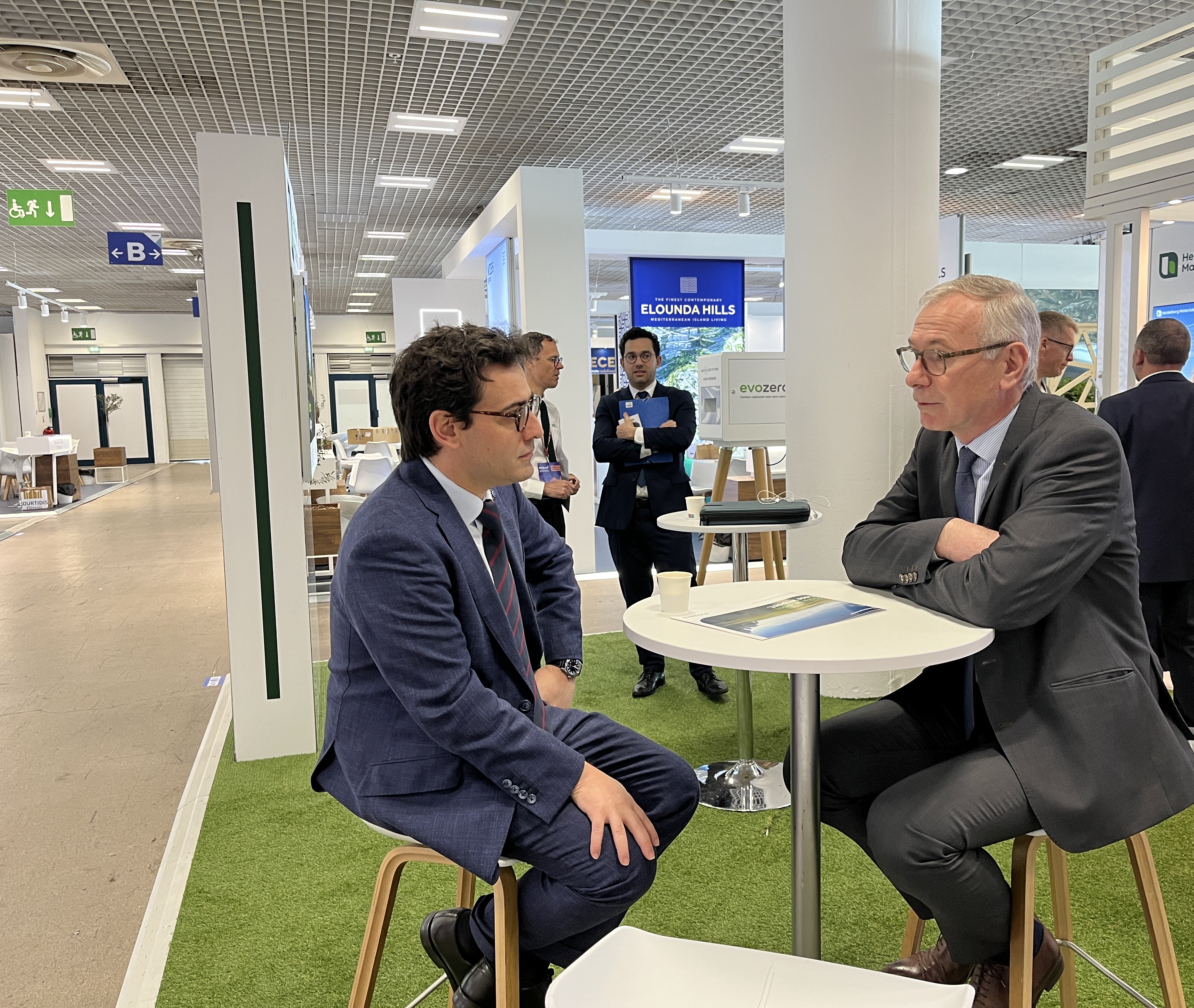 Deux hommes en costume discutent assis à une table dans un espace d'exposition professionnel.