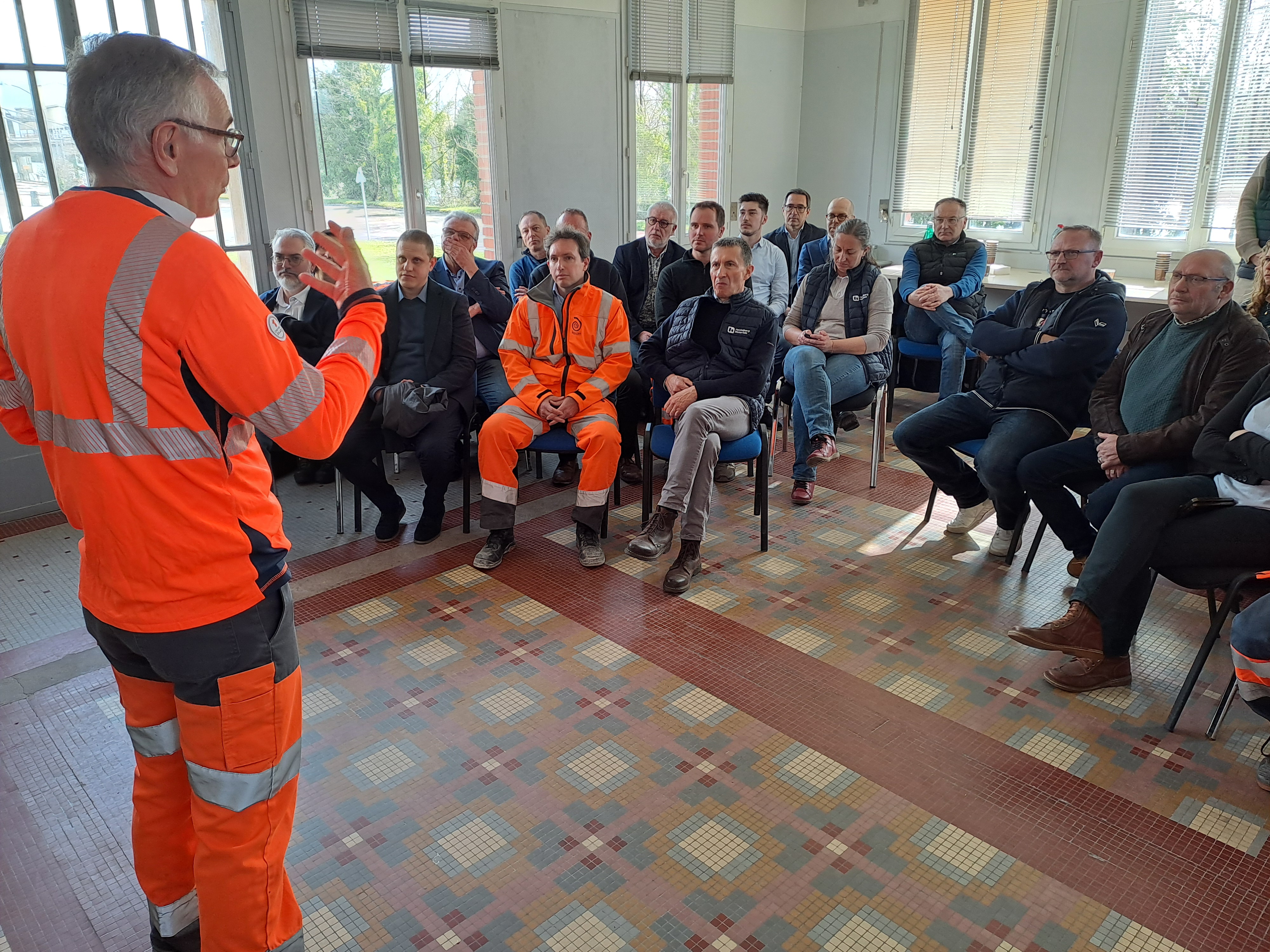 Un groupe de personnes assiste à une présentation dans une salle, avec un intervenant portant une tenue haute visibilité.