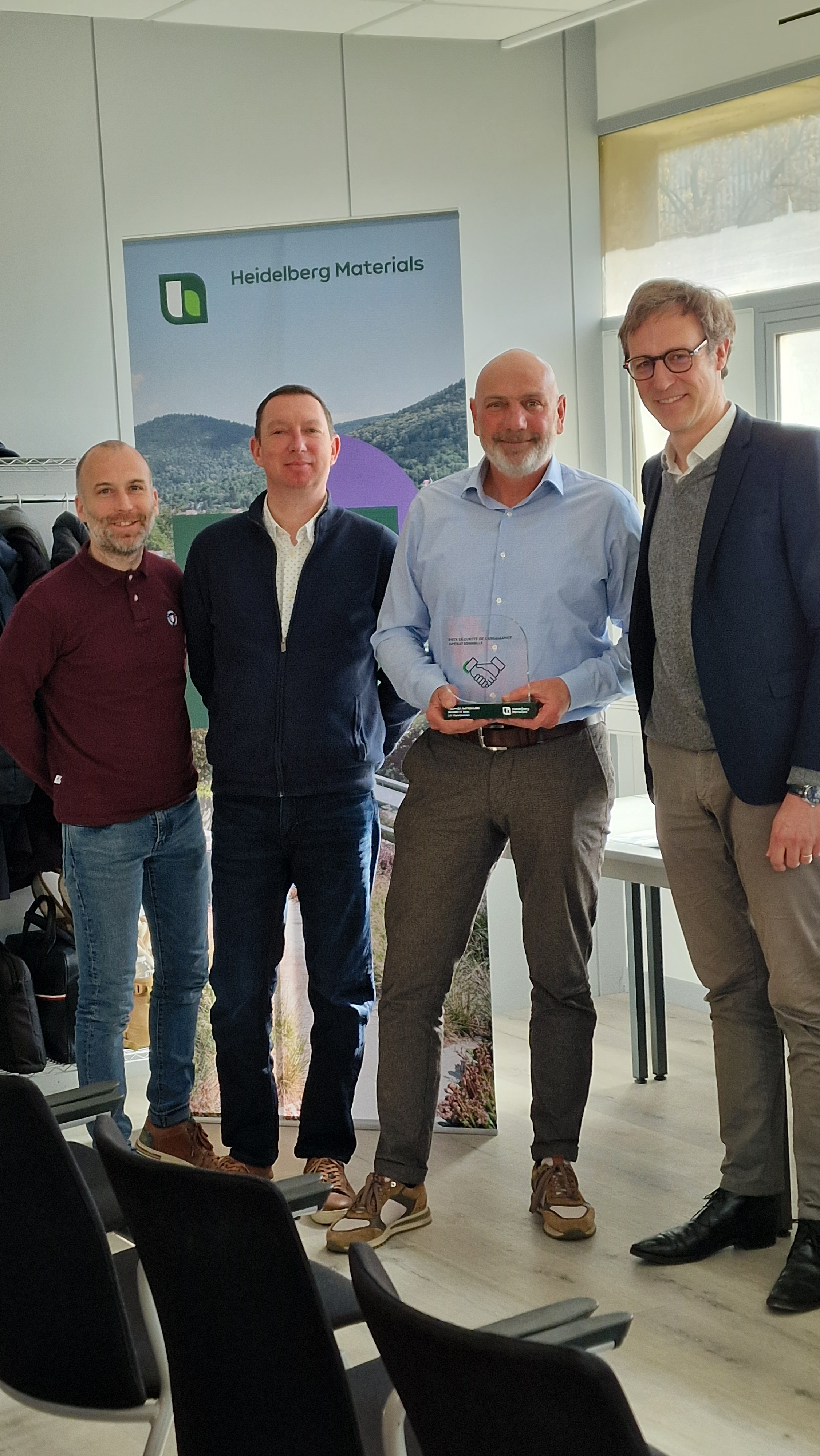 Quatre hommes debout dans une salle de réunion, l'un d'eux tenant un trophée, devant un panneau Heidelberg Materials.