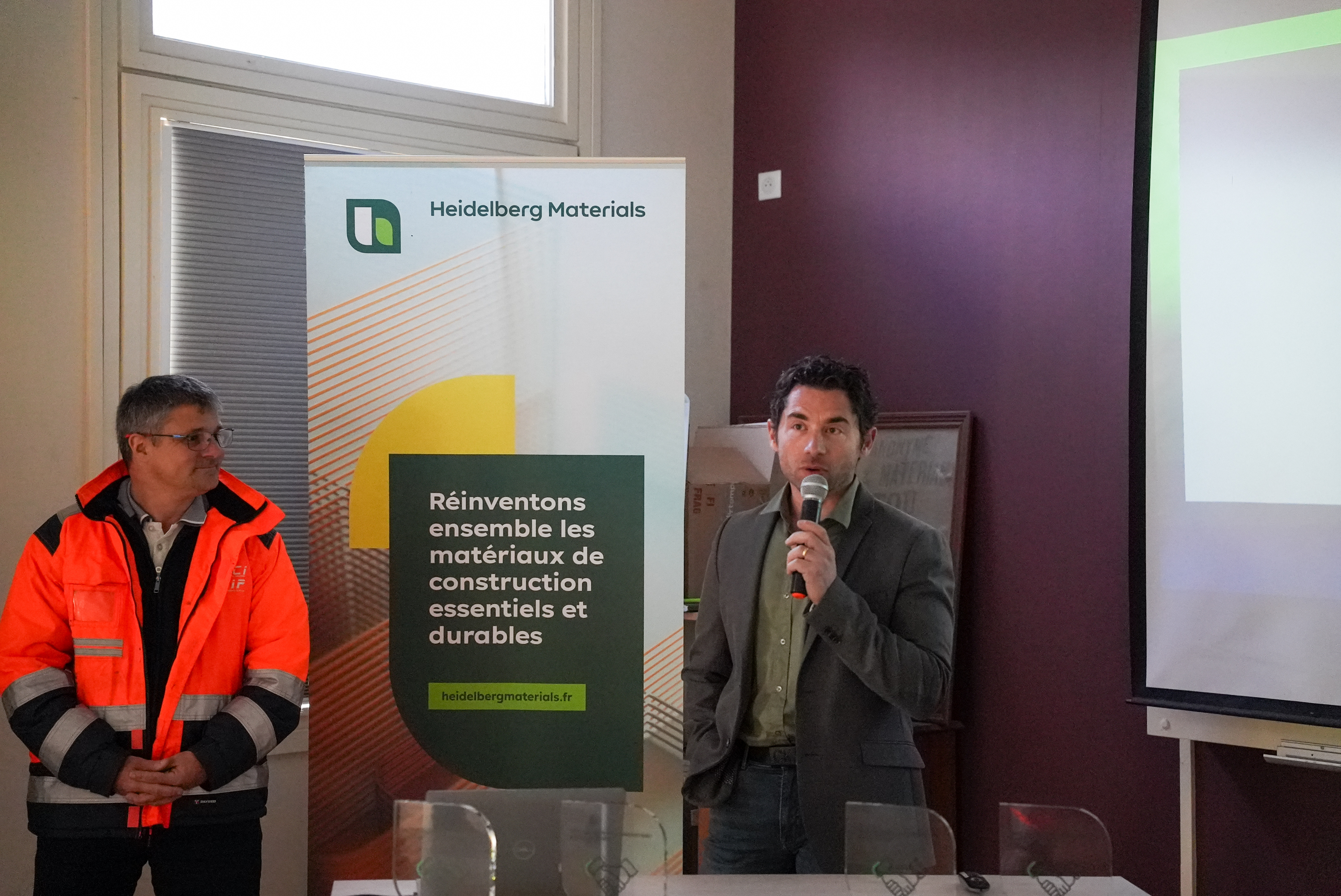 Deux hommes présentent devant un panneau Heidelberg Materials dans un contexte professionnel. 