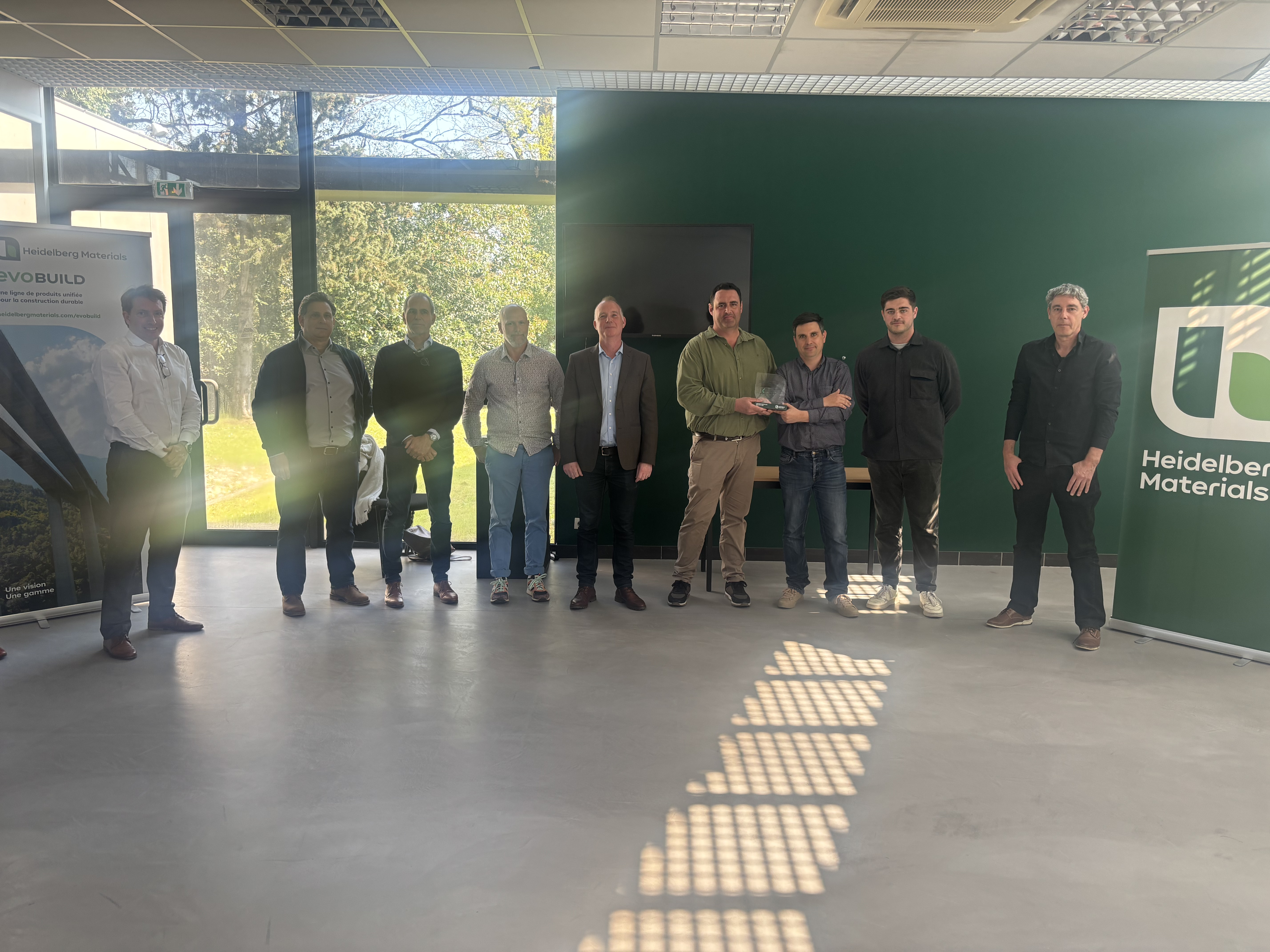 Un groupe de personnes posant dans une salle avec des panneaux et un mur vert, utilisé pour une communication corporative de Heidelberg Materials.