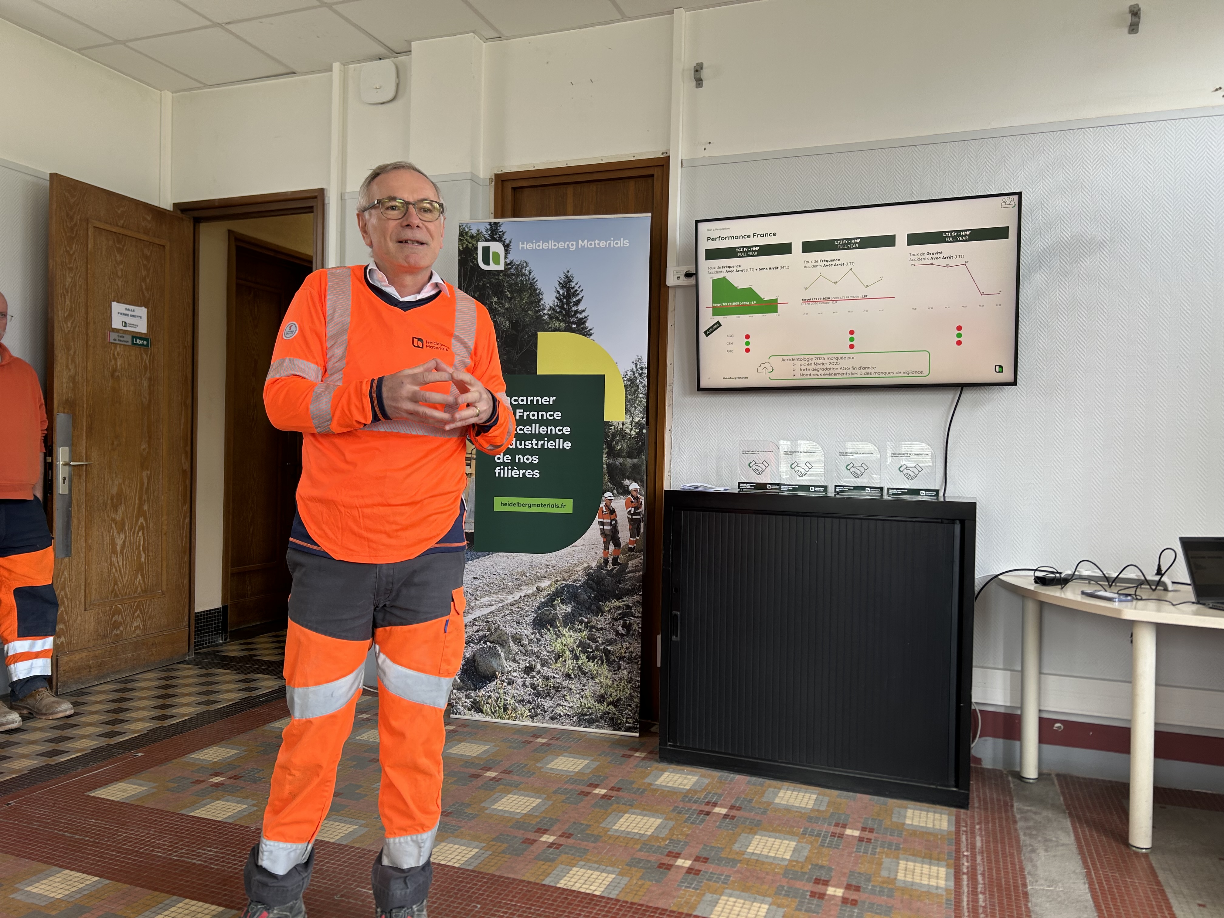 Un homme en tenue de sécurité orange donne une présentation avec un écran affichant des graphiques en arrière-plan.