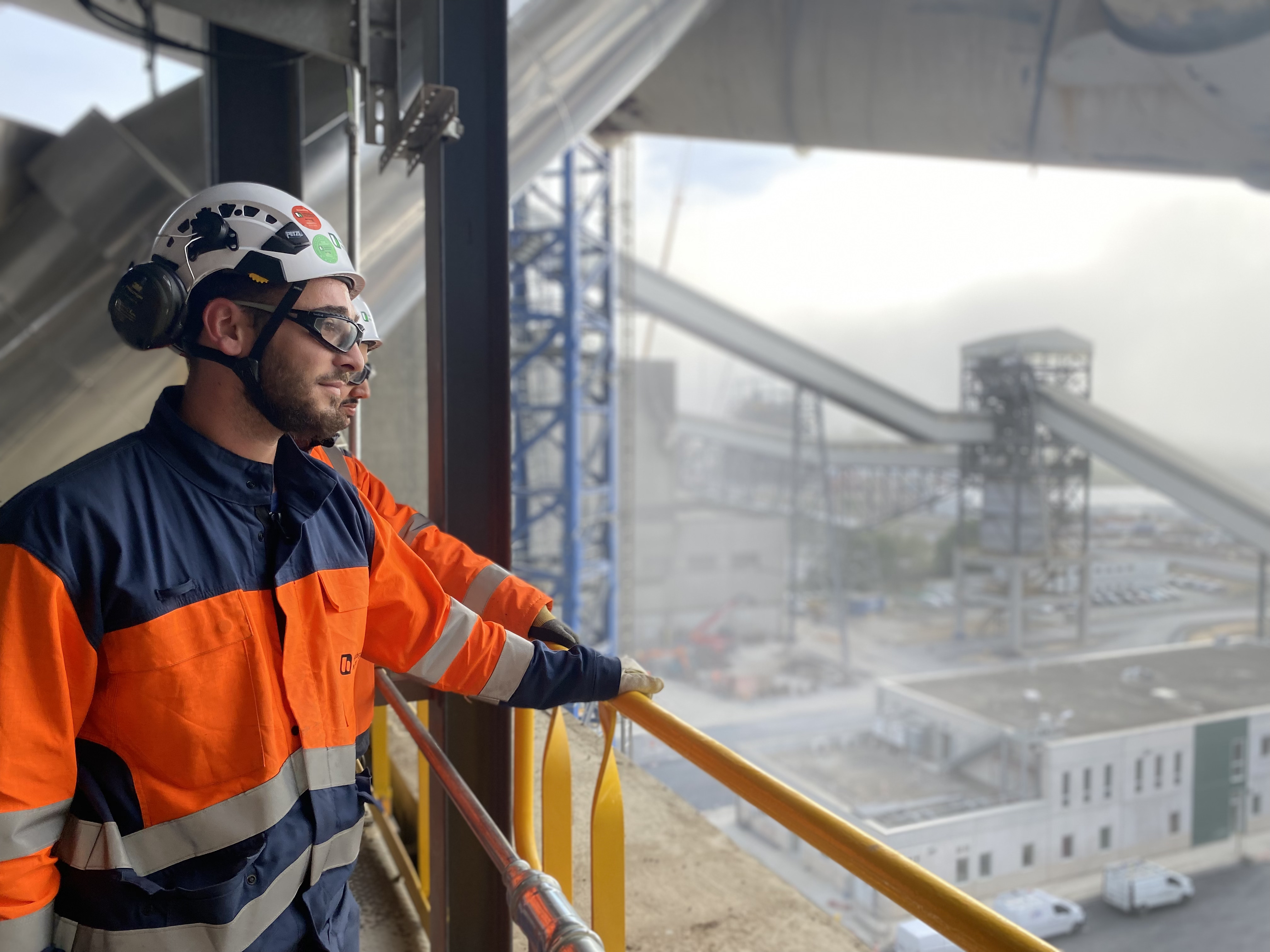 Un employé en tenue de sécurité observe un site industriel avec des convoyeurs et des bâtiments.