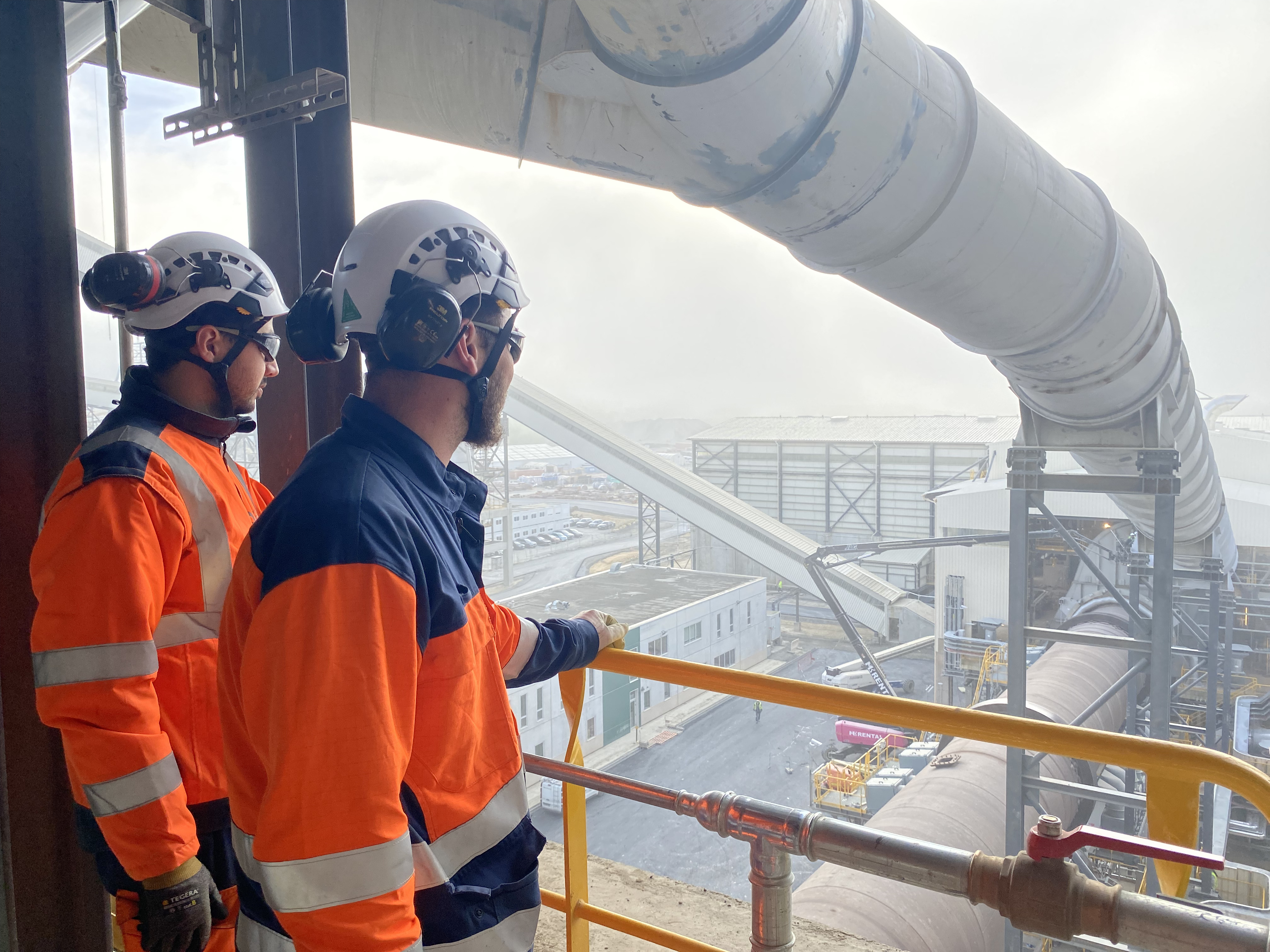 Deux employés en tenue de sécurité observent une installation industrielle avec des convoyeurs.