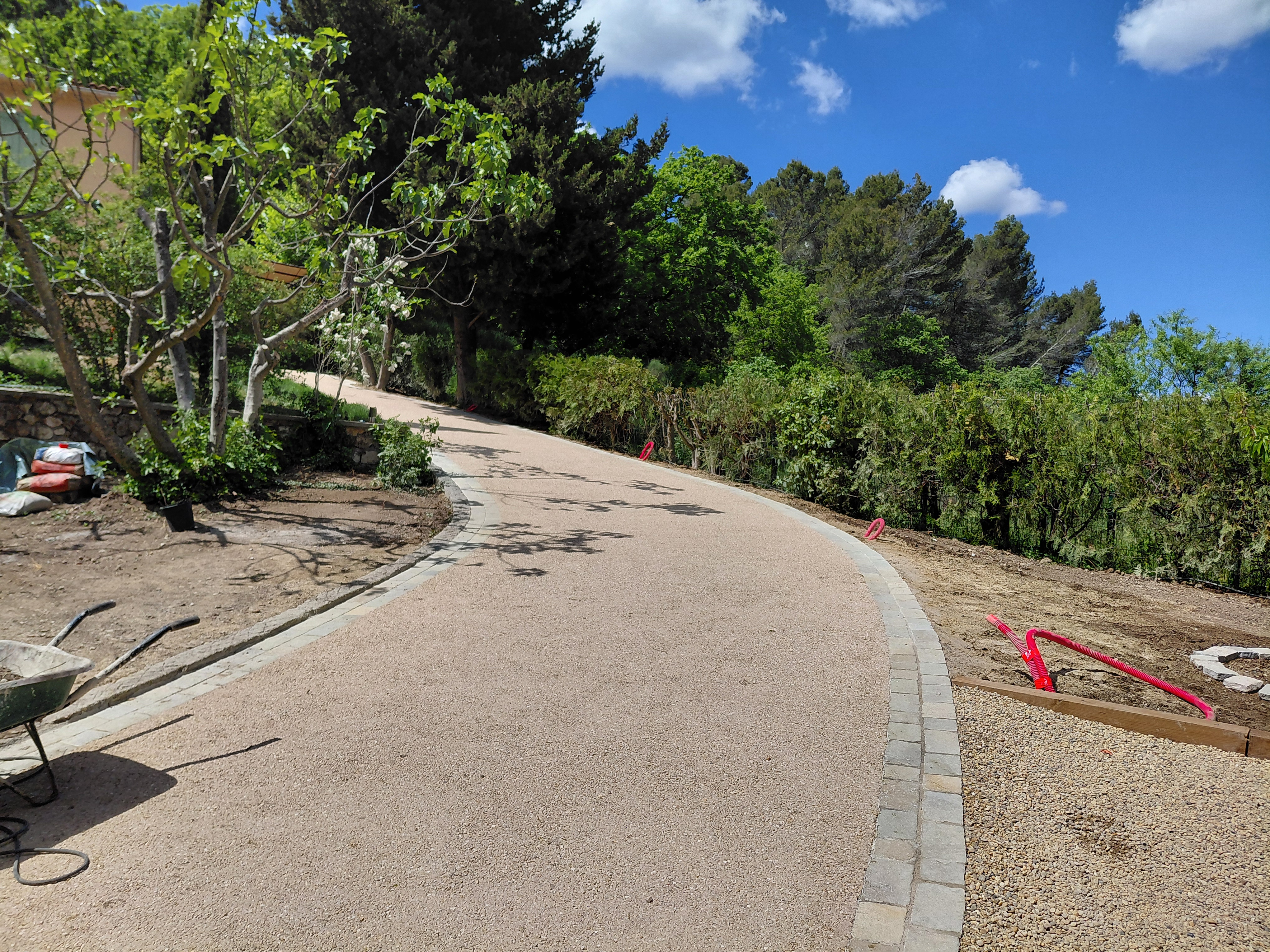 Allée en gravier bordée de végétation et d'arbres, avec des outils de construction visibles à proximité.