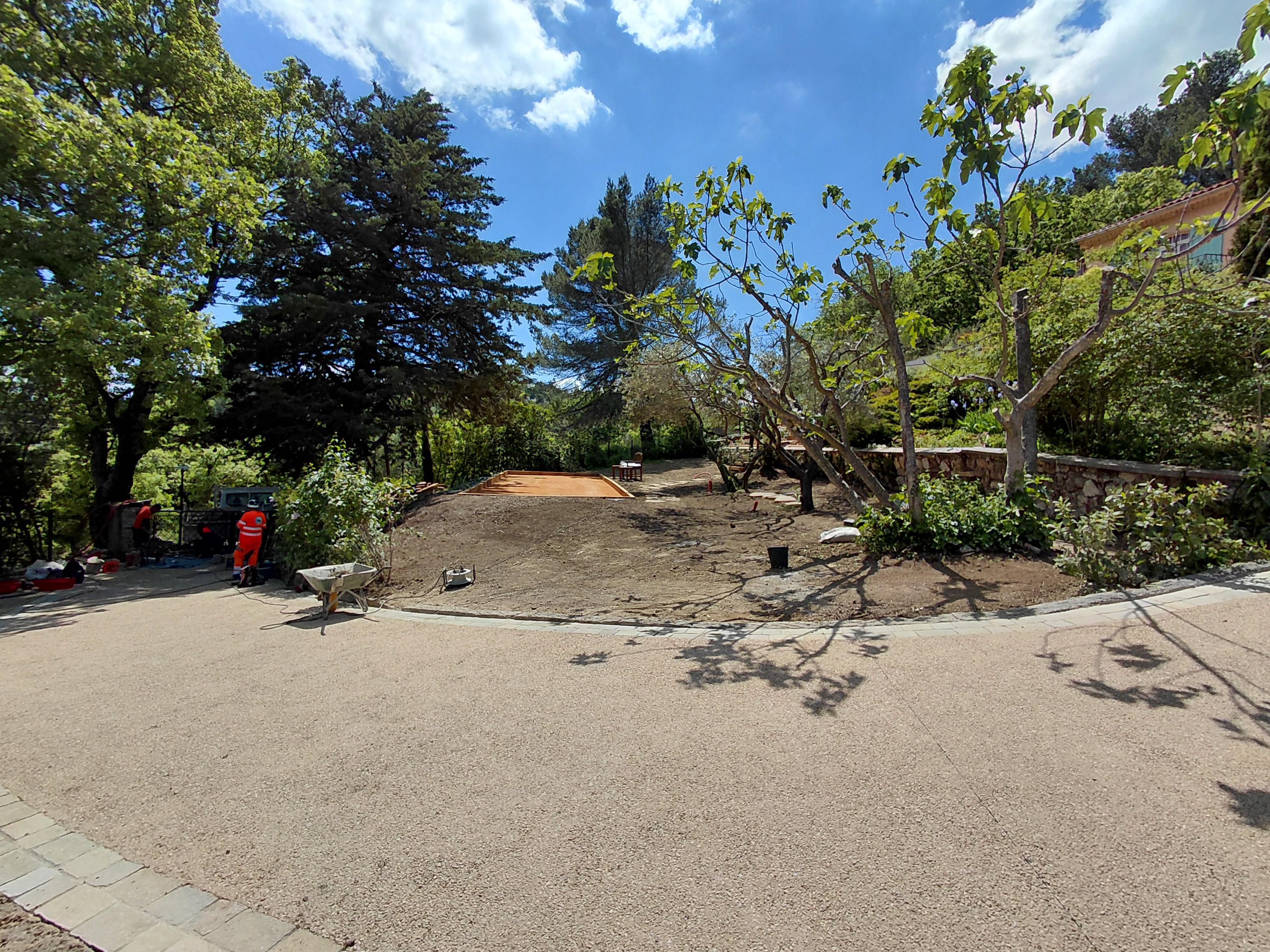 Vue d'un espace extérieur en cours d'aménagement, avec des arbres et des travailleurs visibles à gauche.