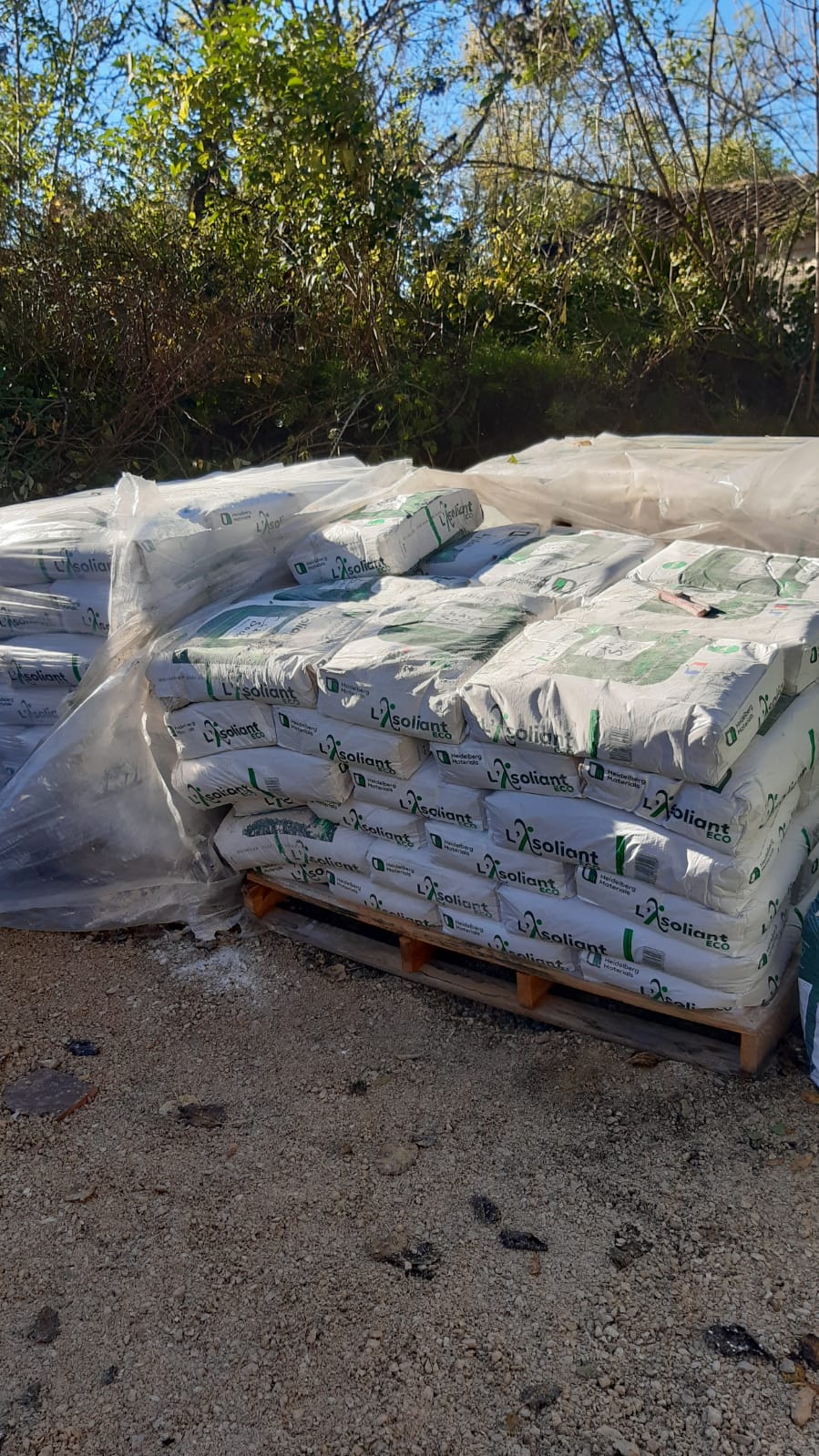 Palette de sacs de matériaux de construction empilés et enveloppés dans un film plastique, entreposée en extérieur.