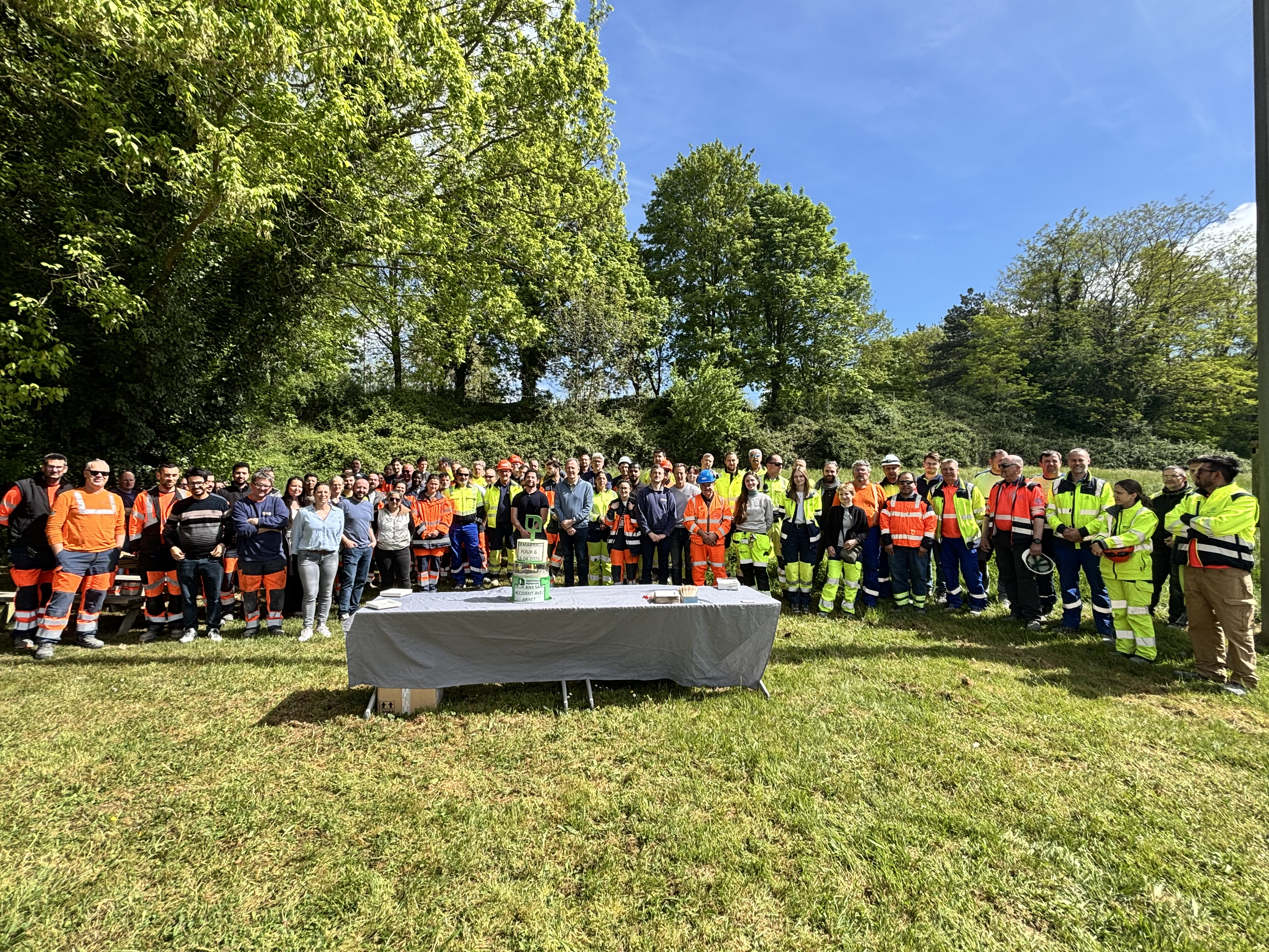 Un grand groupe de personnes en tenue de travail ou décontractée est rassemblé en plein air près d’une table, entouré de verdure.