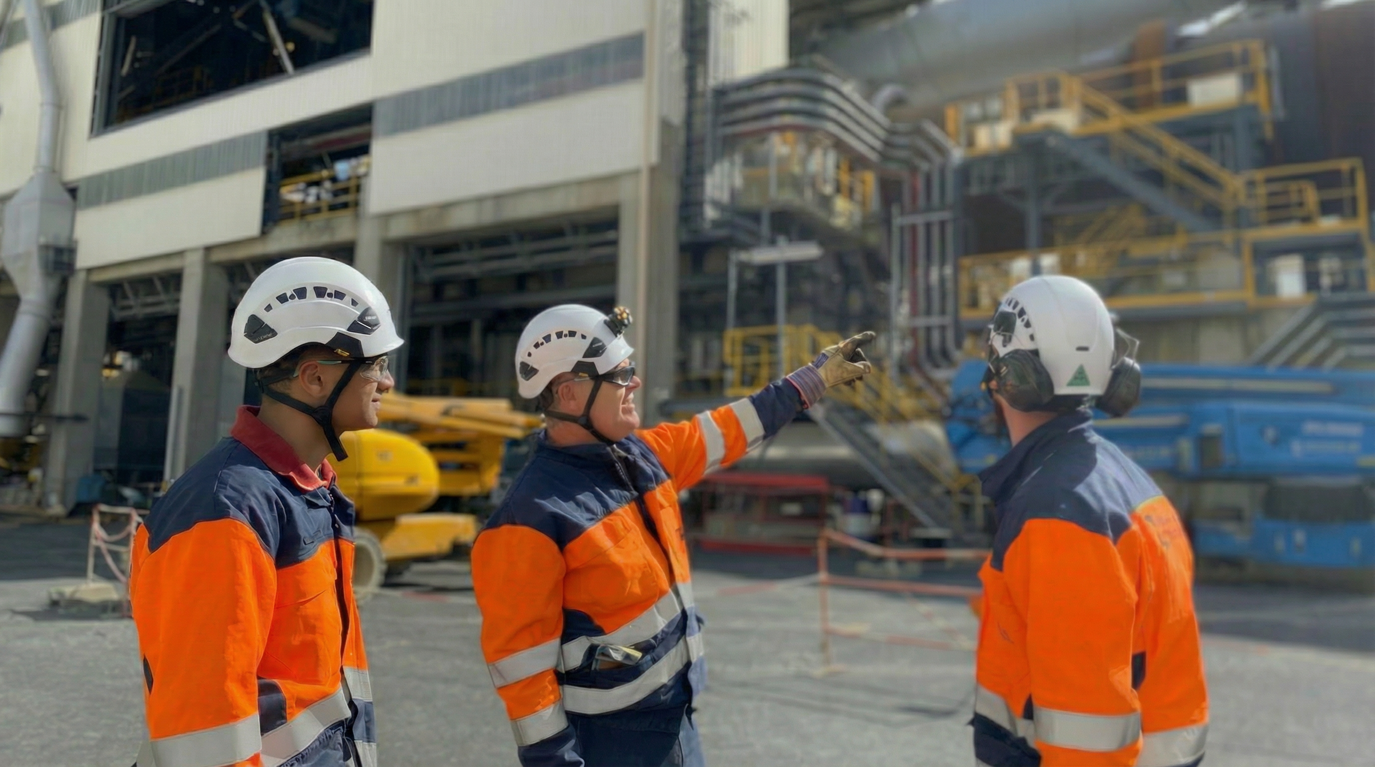 Des employés en tenue de sécurité discutent sur un site industriel avec des équipements de construction en arrière-plan.