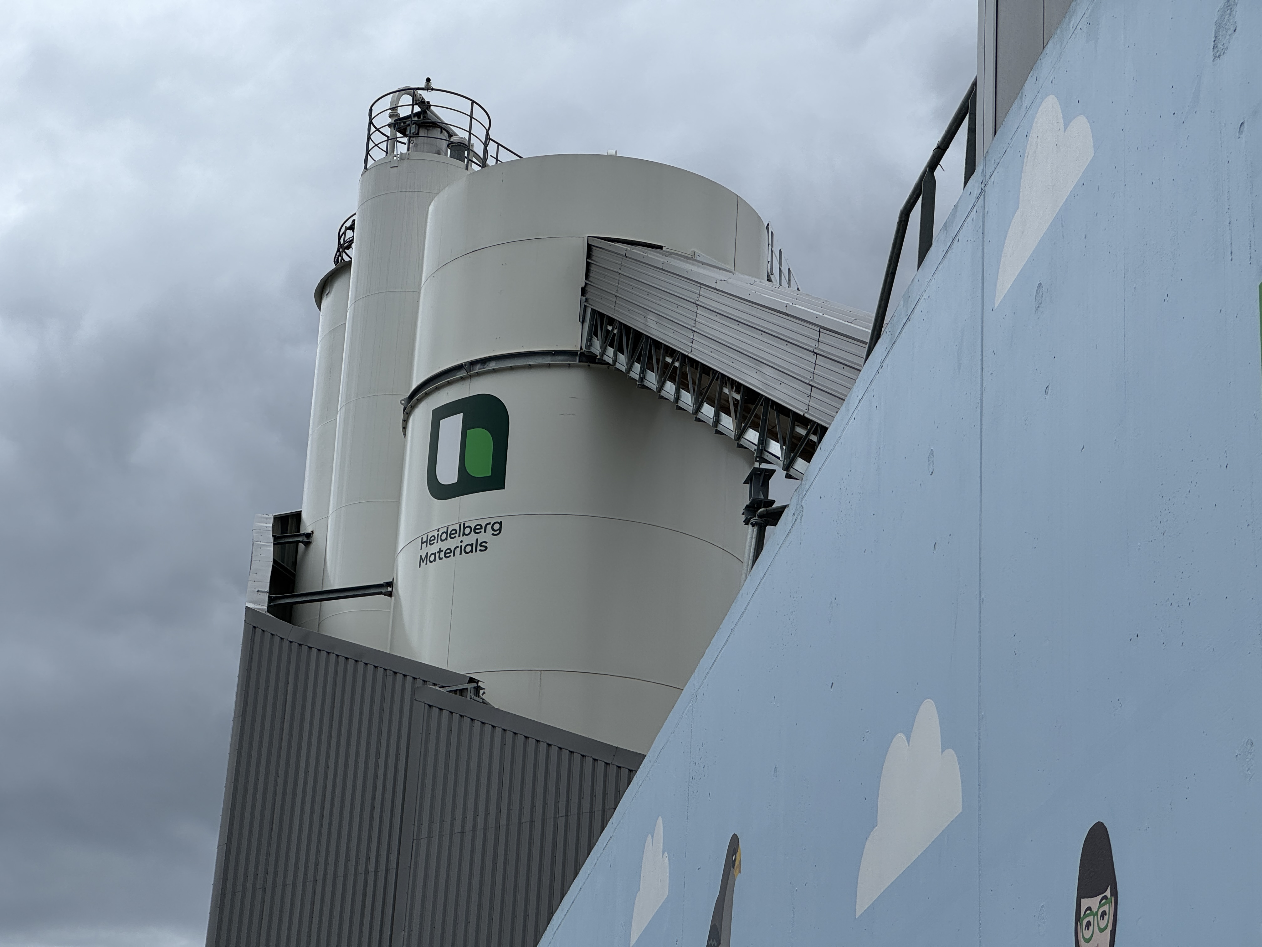 Des silos industriels avec le logo de Heidelberg Materials, visibles derrière un mur décoré de motifs de nuages.