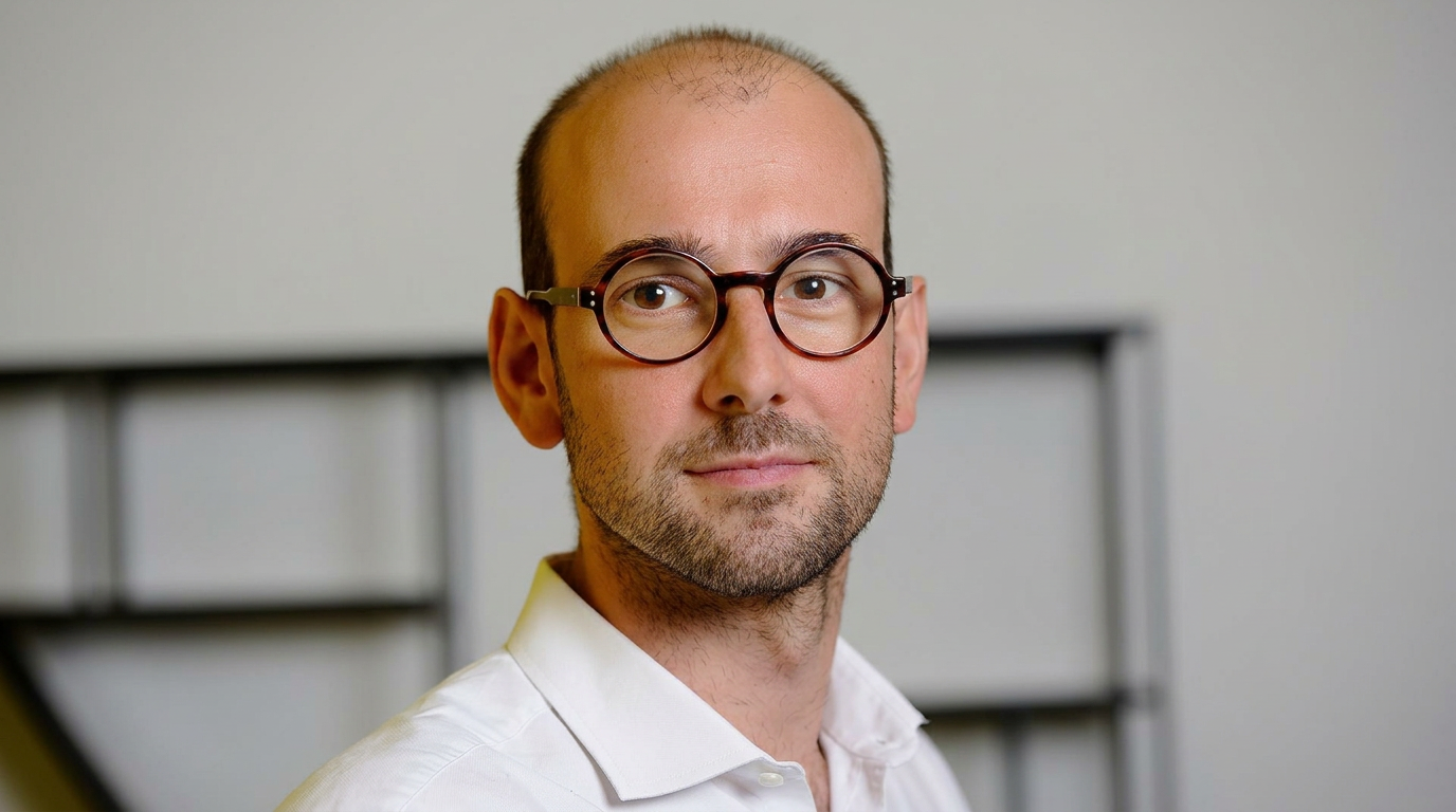 Portrait d'une personne portant des lunettes et une chemise blanche devant un arrière-plan neutre.