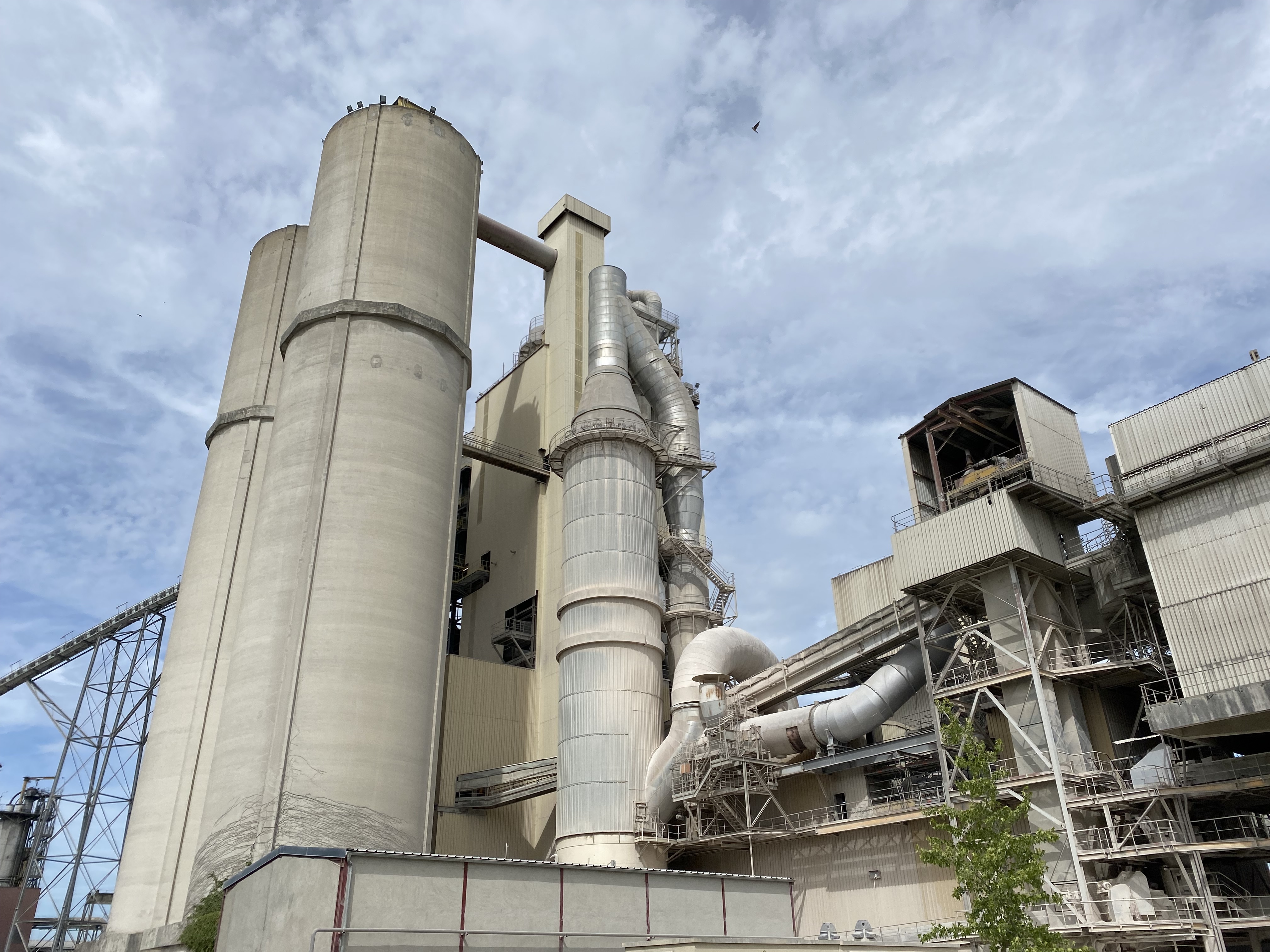 Une grande cimenterie industrielle, avec ses hauts silos et son réseau complexe de canalisations, sous un ciel nuageux.