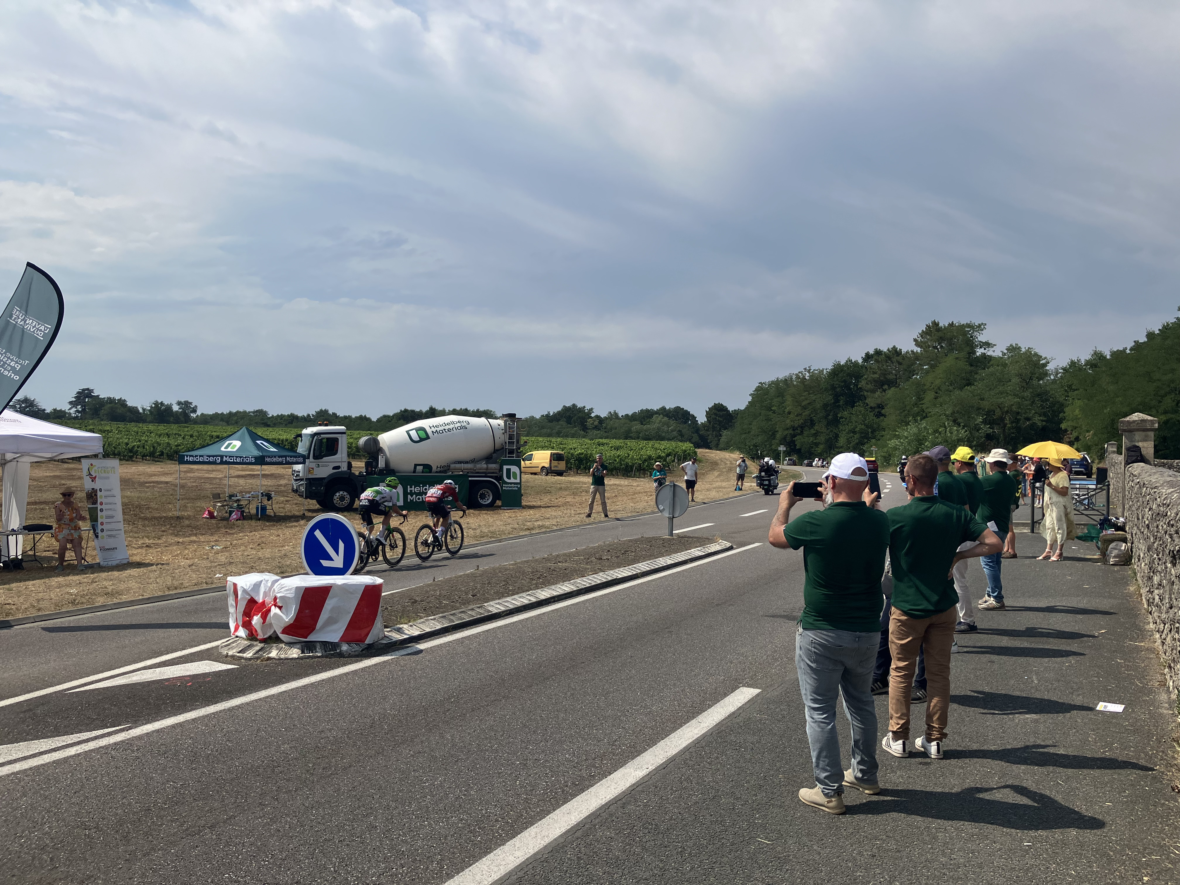 Des spectateurs assistent au passage d'échappés du Tour de France 2026 sur la D11 à Illats. Au second plan, la fan zone et le camion-toupie Heidelberg Materials.