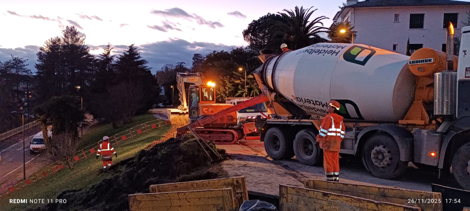 Un camion-toupie décharge du béton sur un site de construction au crépuscule, avec des ouvriers en tenue de sécurité.