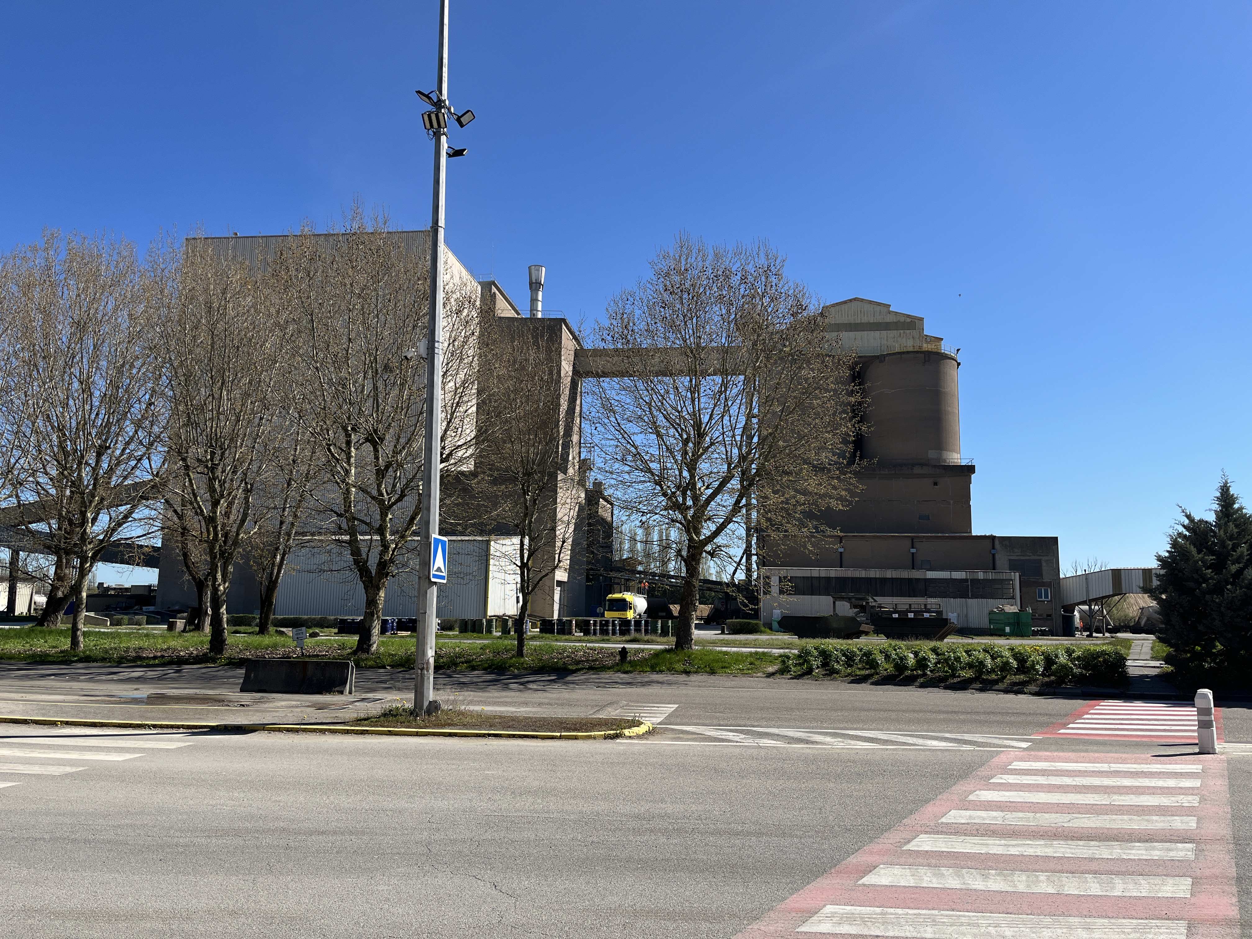 Un grand bâtiment industriel avec des constructions adjacentes, entouré d'arbres, vu depuis l'autre côté de la rue.