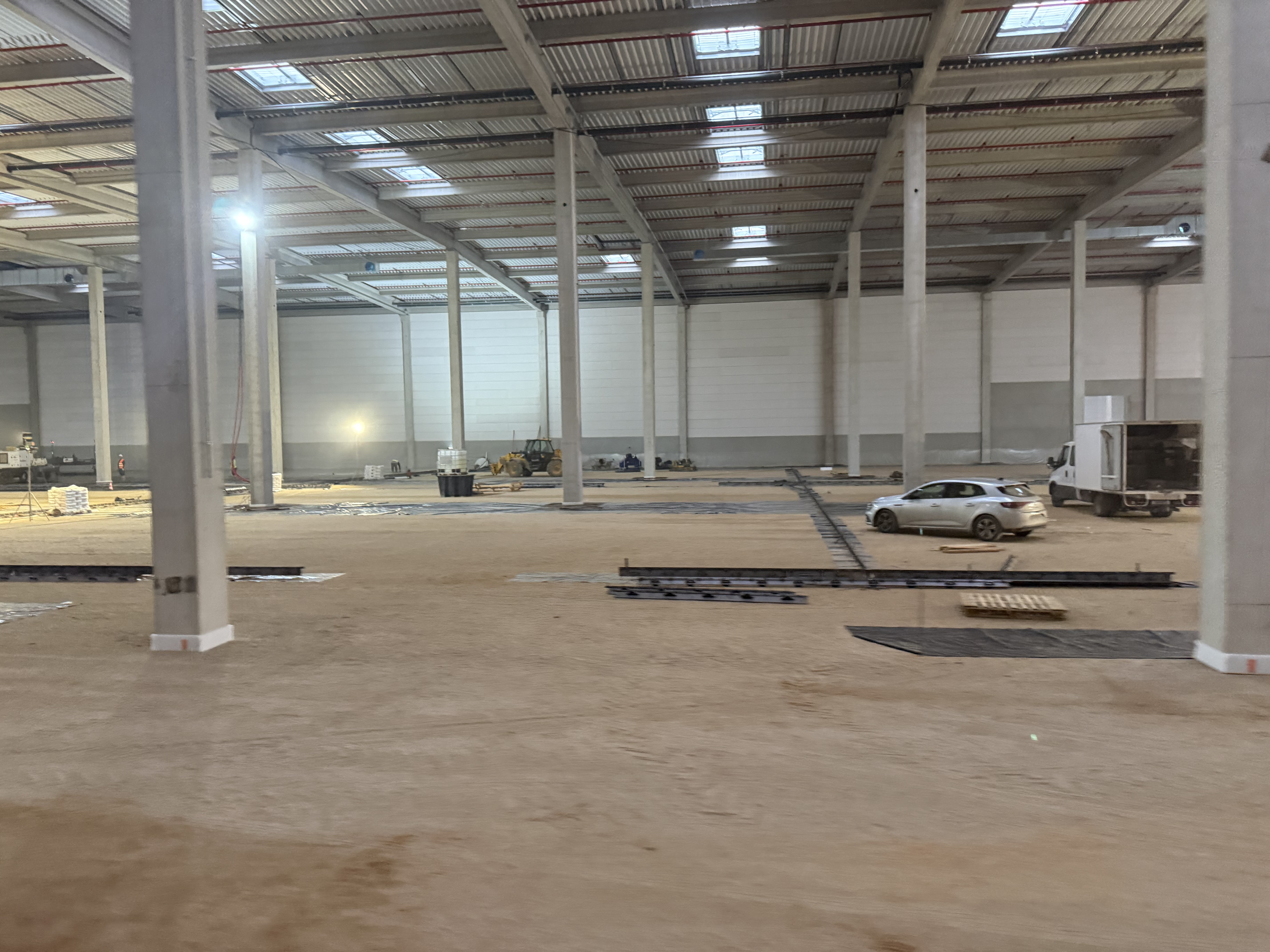 Vue intérieure du futur hub logistique CULTURA à Ferrières-en-Gâtinais. Les travaux de préparation des dallages industriels sont en cours au sein d'un vaste entrepôt doté d'une structure en béton et acier, avec plusieurs véhicules et équipements de chantier visibles.