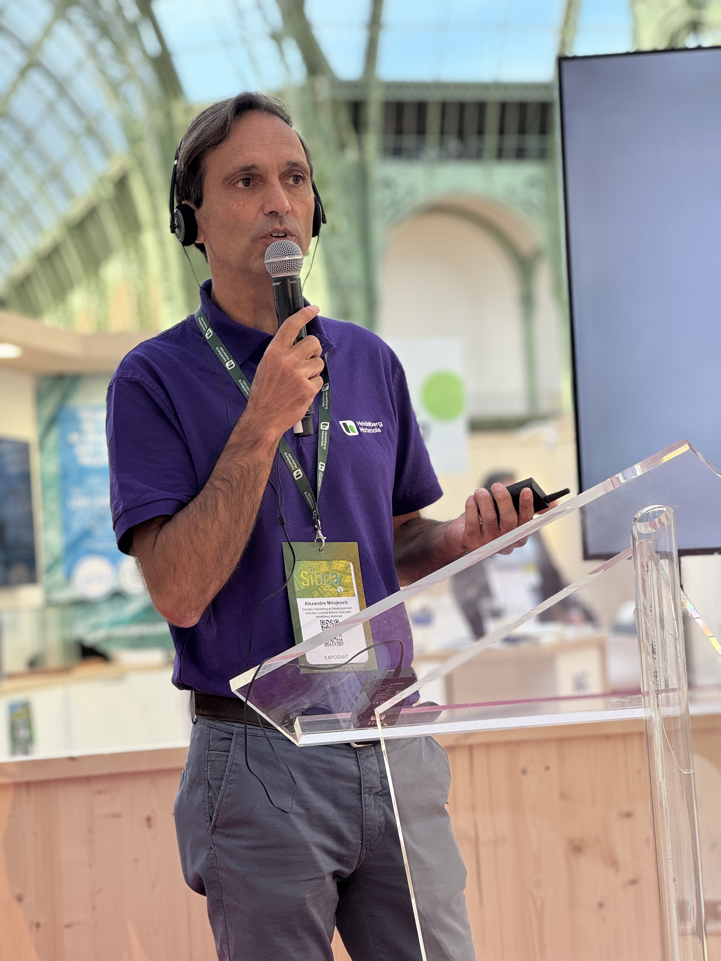Un intervenant en chemise violette et casque-micro parle lors d'une présentation dans un lieu lumineux avec une architecture en verre et métal.