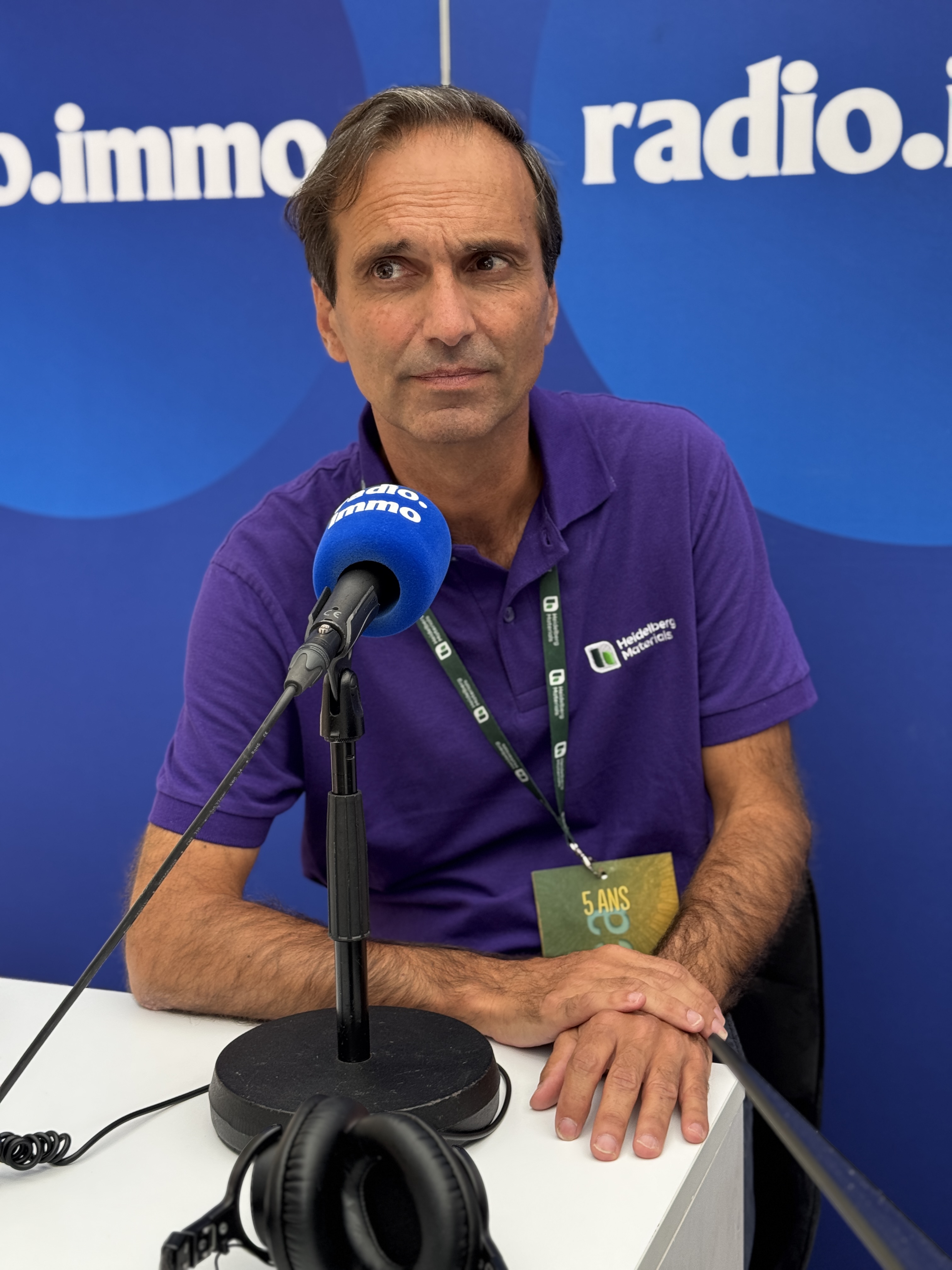 Un homme assis devant un microphone dans un studio, portant un polo violet avec un logo d'entreprise.