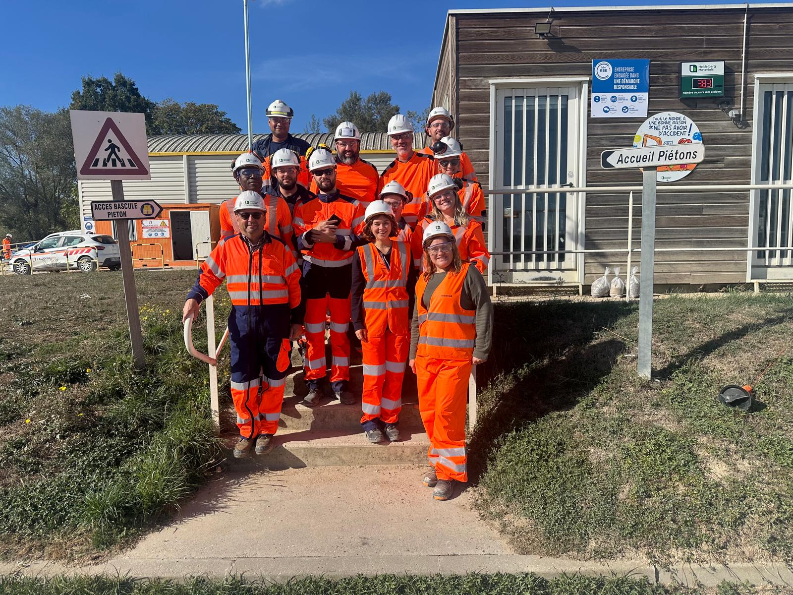 Un groupe de travailleurs en tenue de sécurité orange pose devant un bâtiment sur un site industriel.