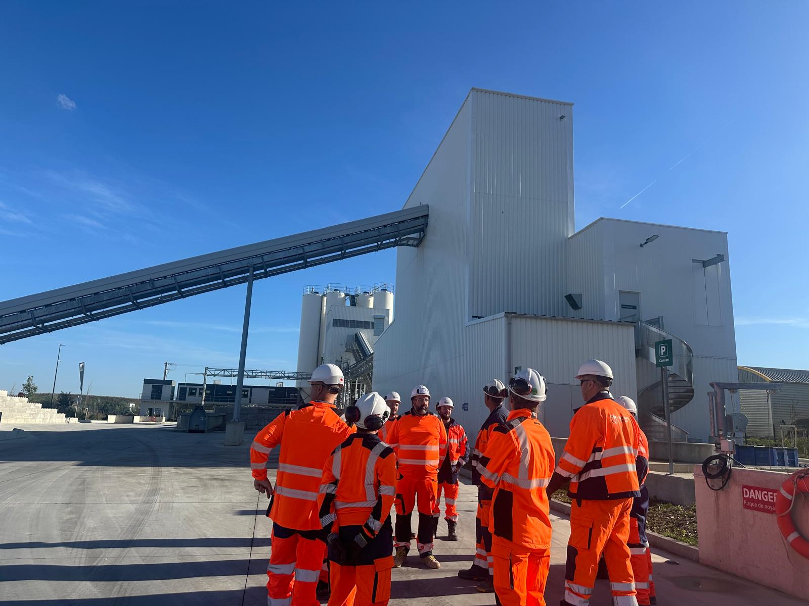 Un groupe de personnes en tenue de sécurité orange se tient devant un bâtiment industriel sur un site de construction.